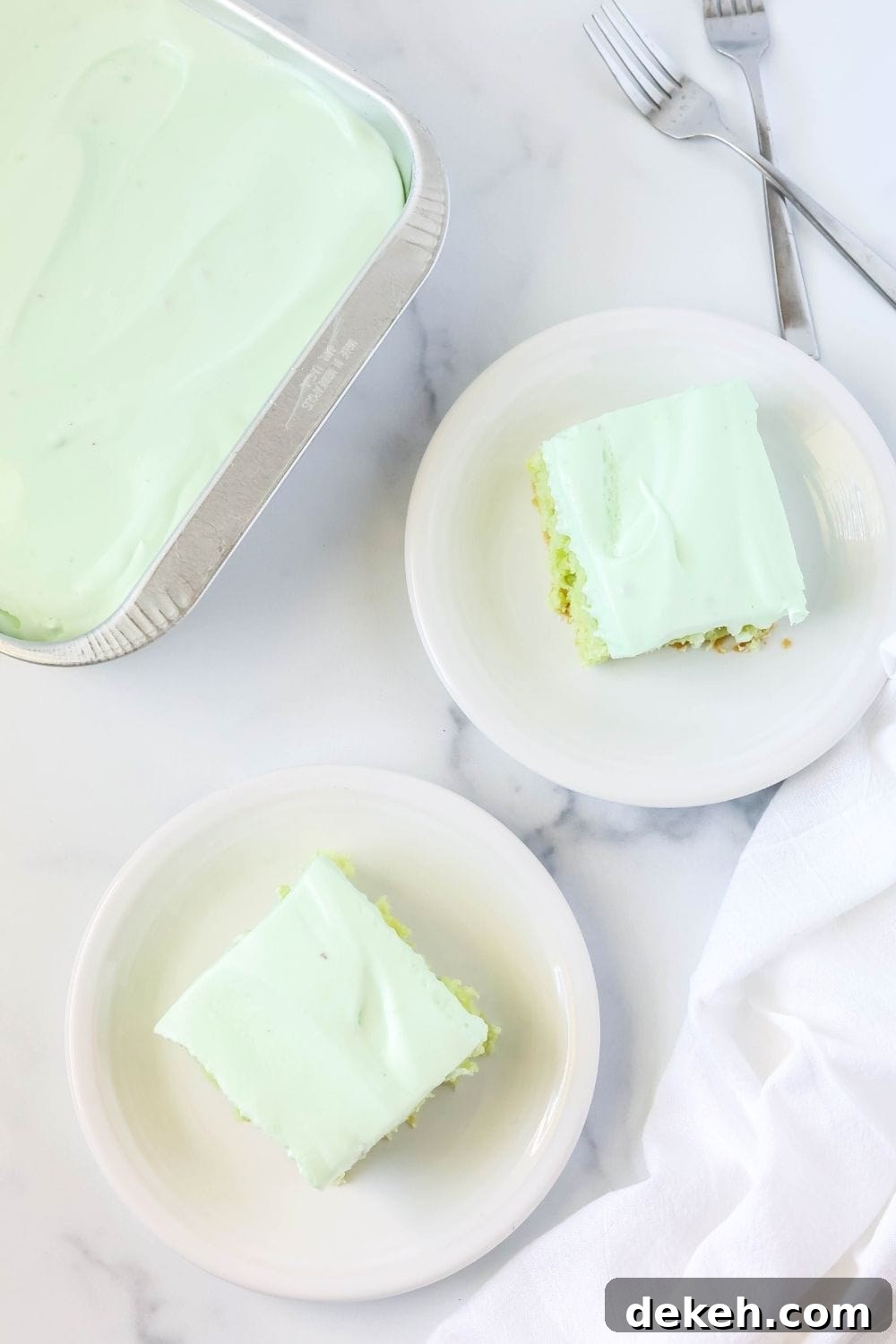 overhead view of two white plates serving pieces of pineapple pistachio cake. The pan of remaining cake is next to the plates, along with two forks.