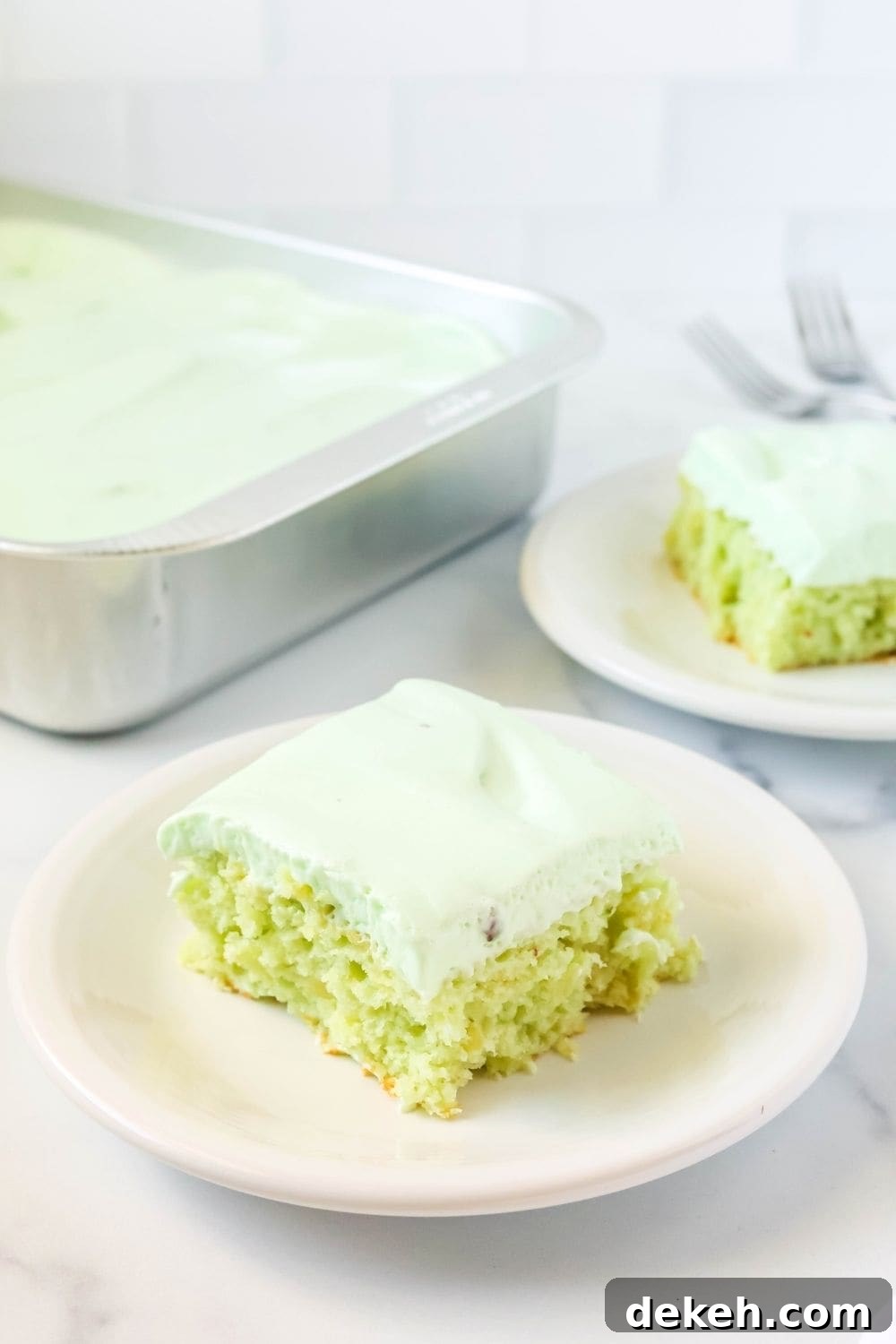 a slice of pistachio pudding and pineapple cake on a white plate, with another slice and the remaining pan of cake in the background.
