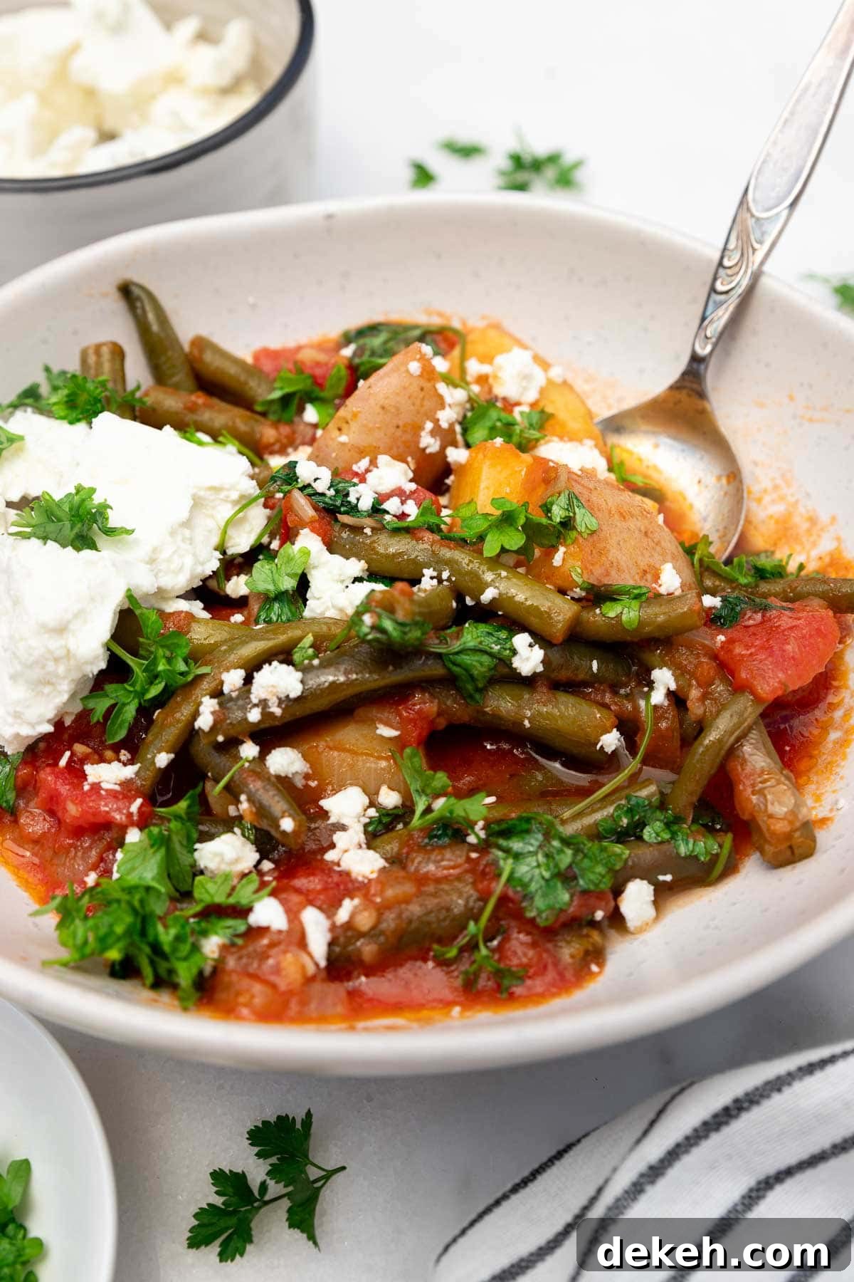 Closeup of a bowl of fasolakia or Greek green beans with a spoon in and feta cheese on top, with ingredients and a tea towel at the side.