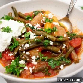 Closeup of a bowl of fasolakia or Greek green beans with a spoon in and feta cheese on top, with ingredients and a tea towel at the side.
