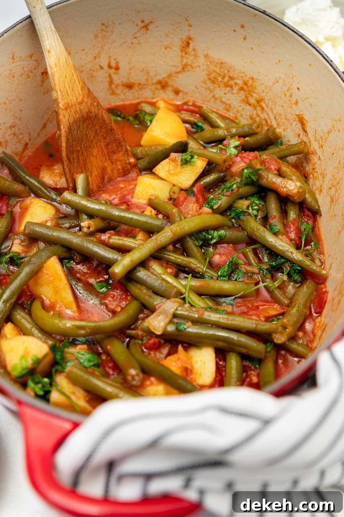 Closeup of a pan of cooked Greek green beans or fasolakia with a wooden spoon in it and a white and black tea towel at the front.