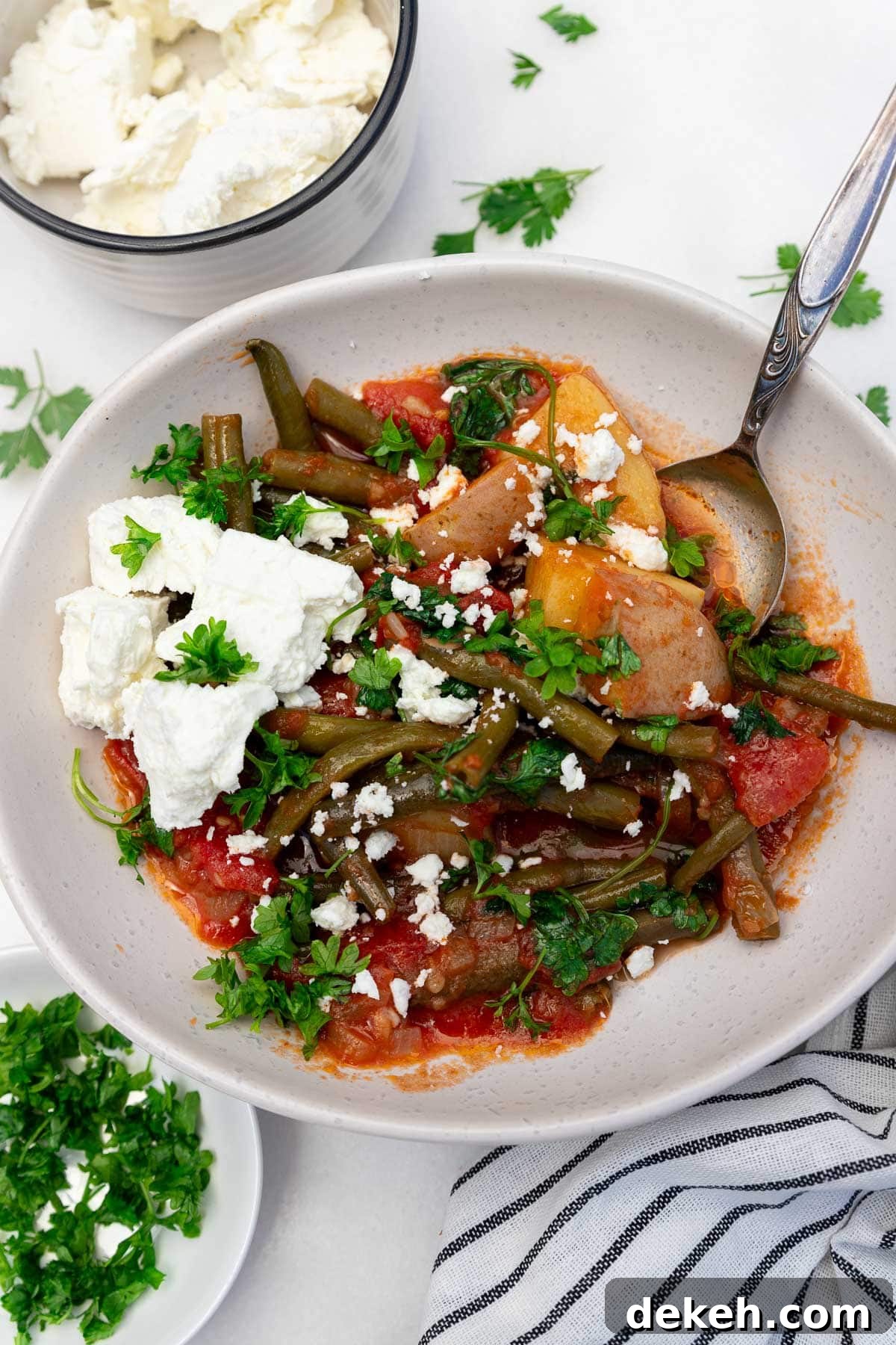 Overhead view of a bowl of fasolakia or Greek green beans with a spoon in and feta cheese on top, with ingredients and a tea towel at the side.