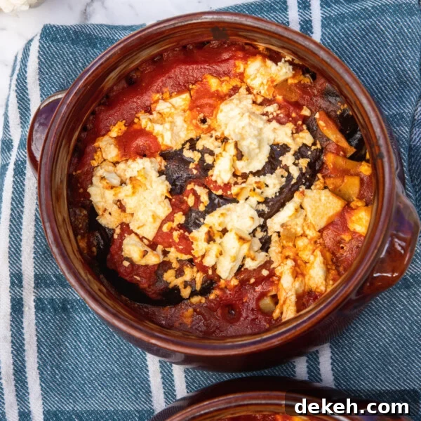 2 brown ceramic dishes of baked eggplant with tomato and feta on a blue and white tea towel and marble background.