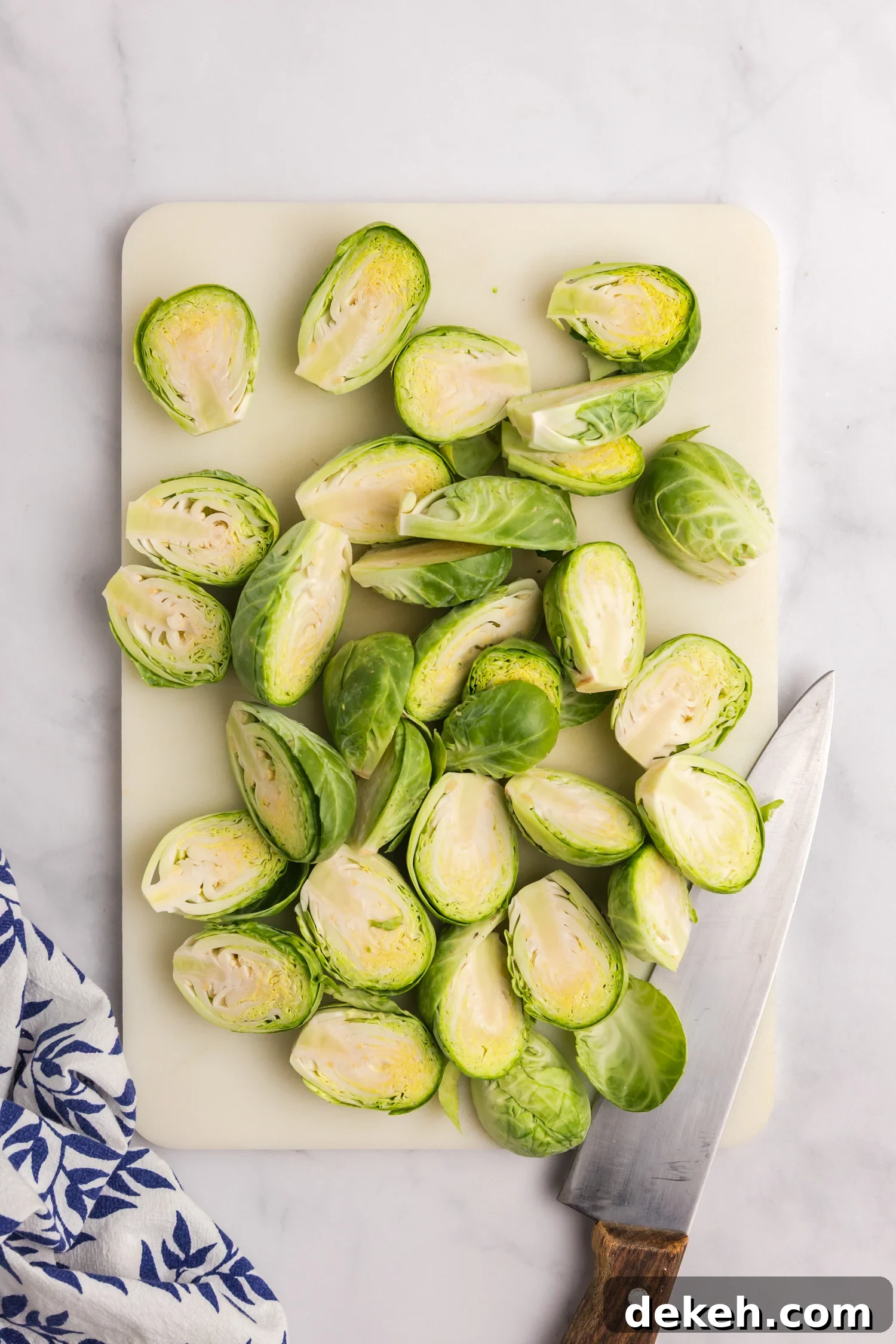 Fresh Brussels sprouts cut in half on a white cutting board with a knife placed to the side.