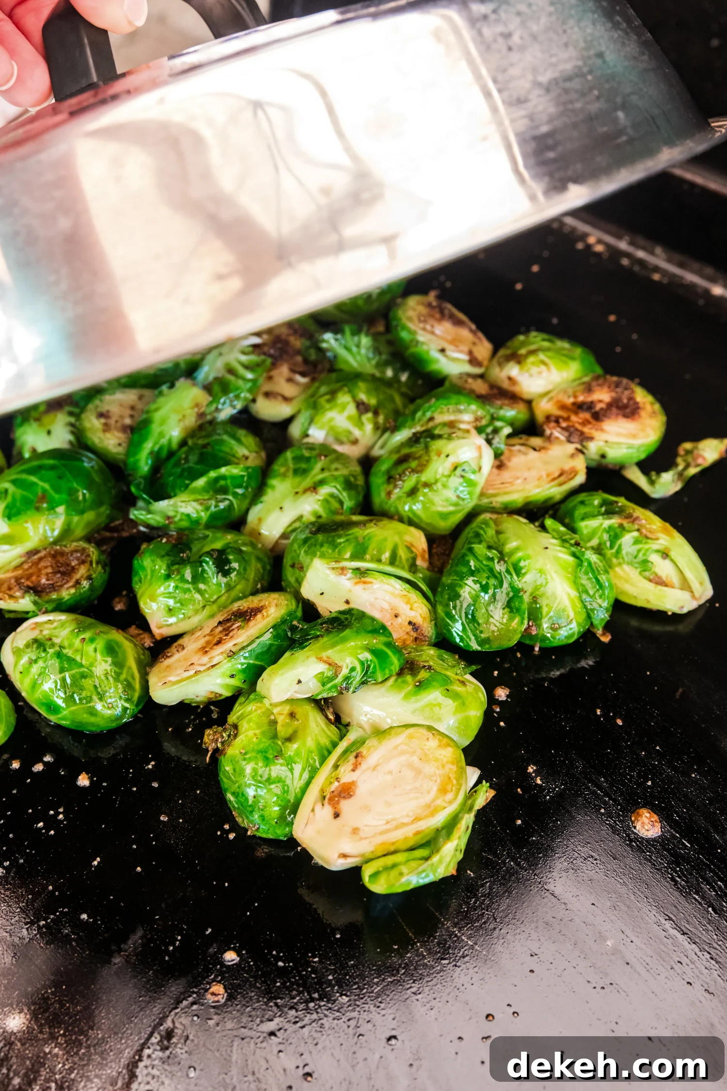 A hand holding a lid over Brussels sprouts on a griddle, allowing them to steam while cooking.