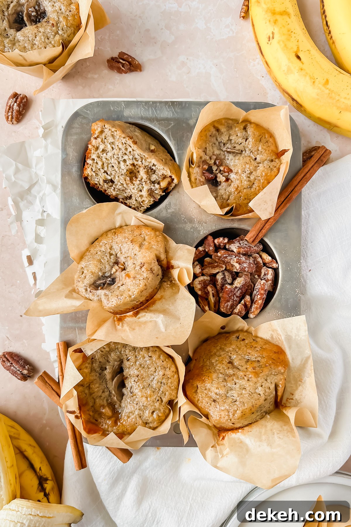 baked greek yogurt banana muffins baked in parchment liners arranged in a 6 muffin muffin tin with pecans, cinnamon sticks, and bananas surrounding.