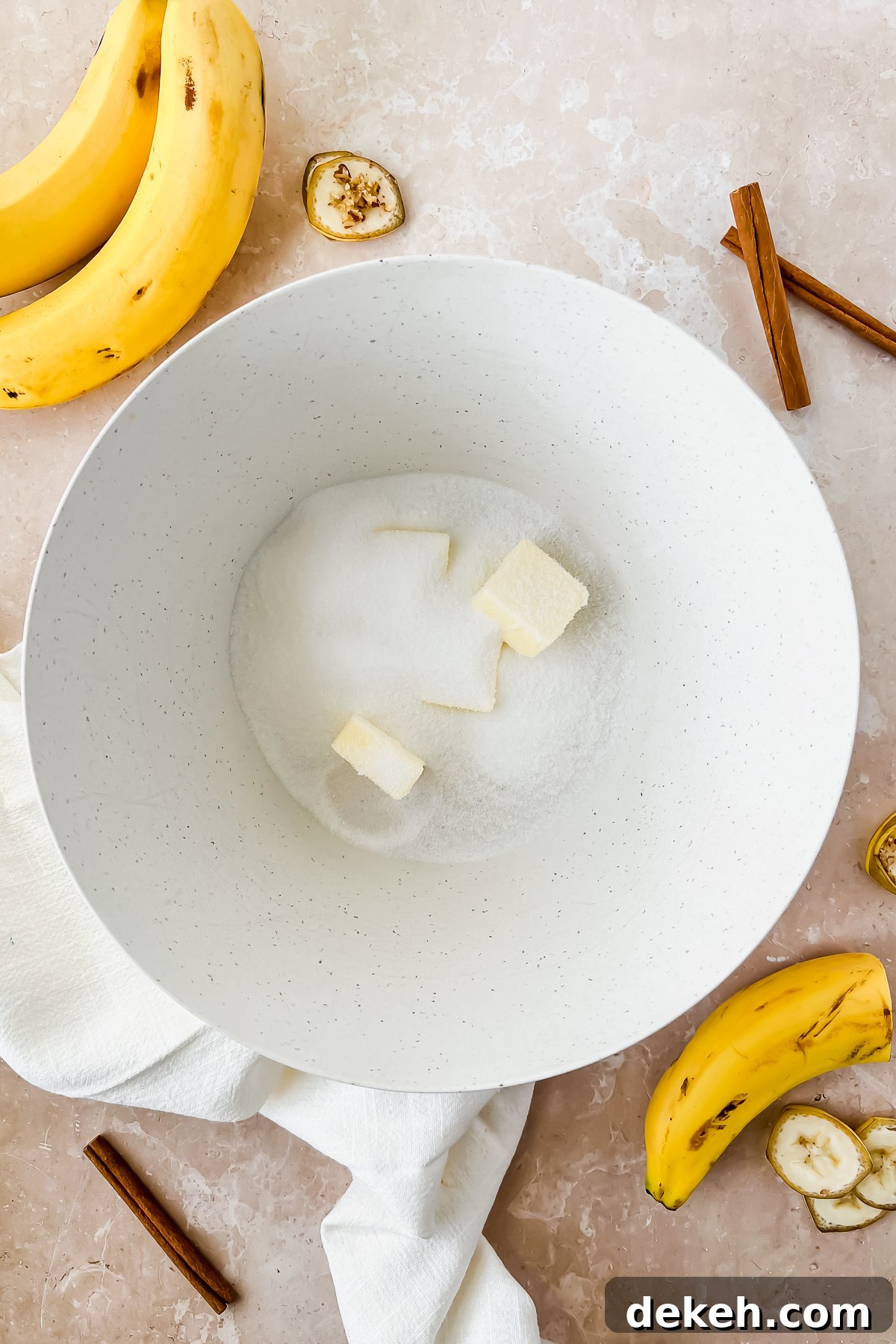 slices of butter and sugar in white mixing bowl beside bananas.