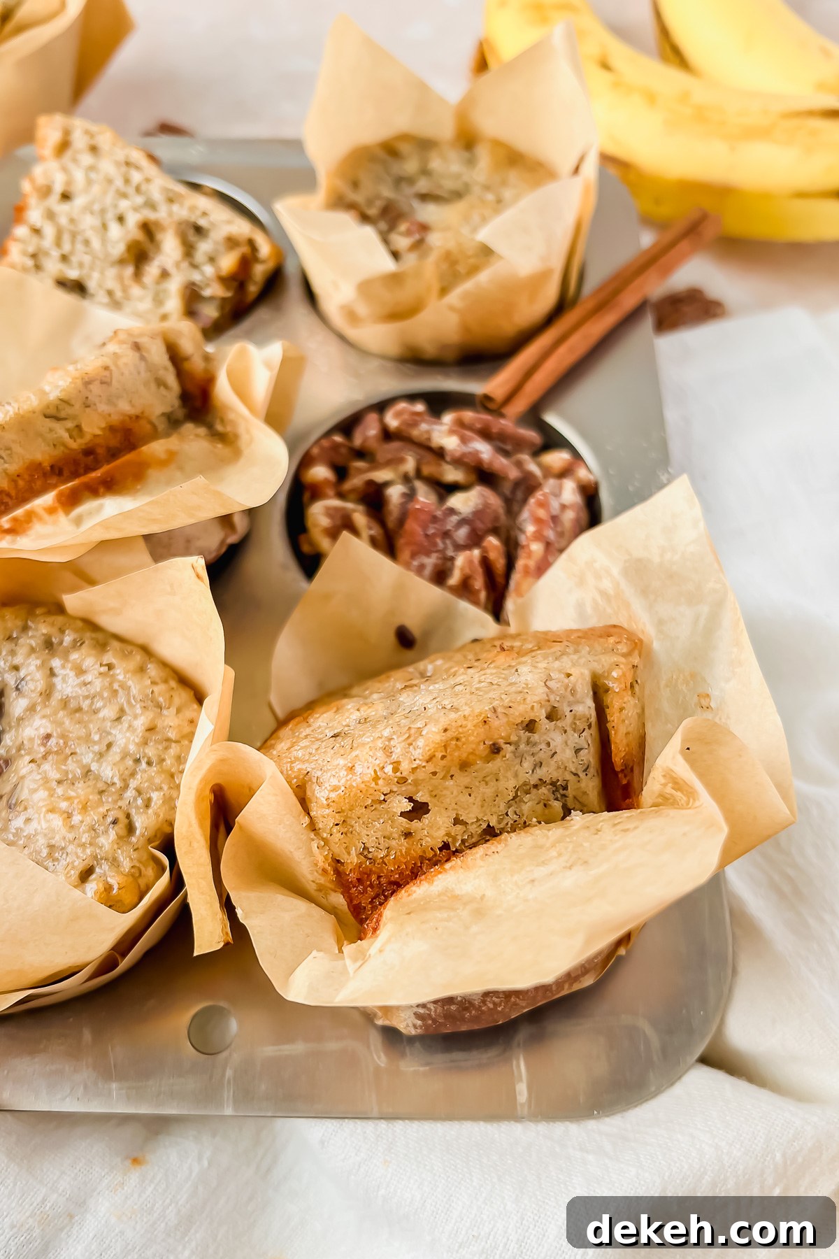 greek yogurt banana muffin in parchment liner in silver baking tin with additional muffins and pecans in background.