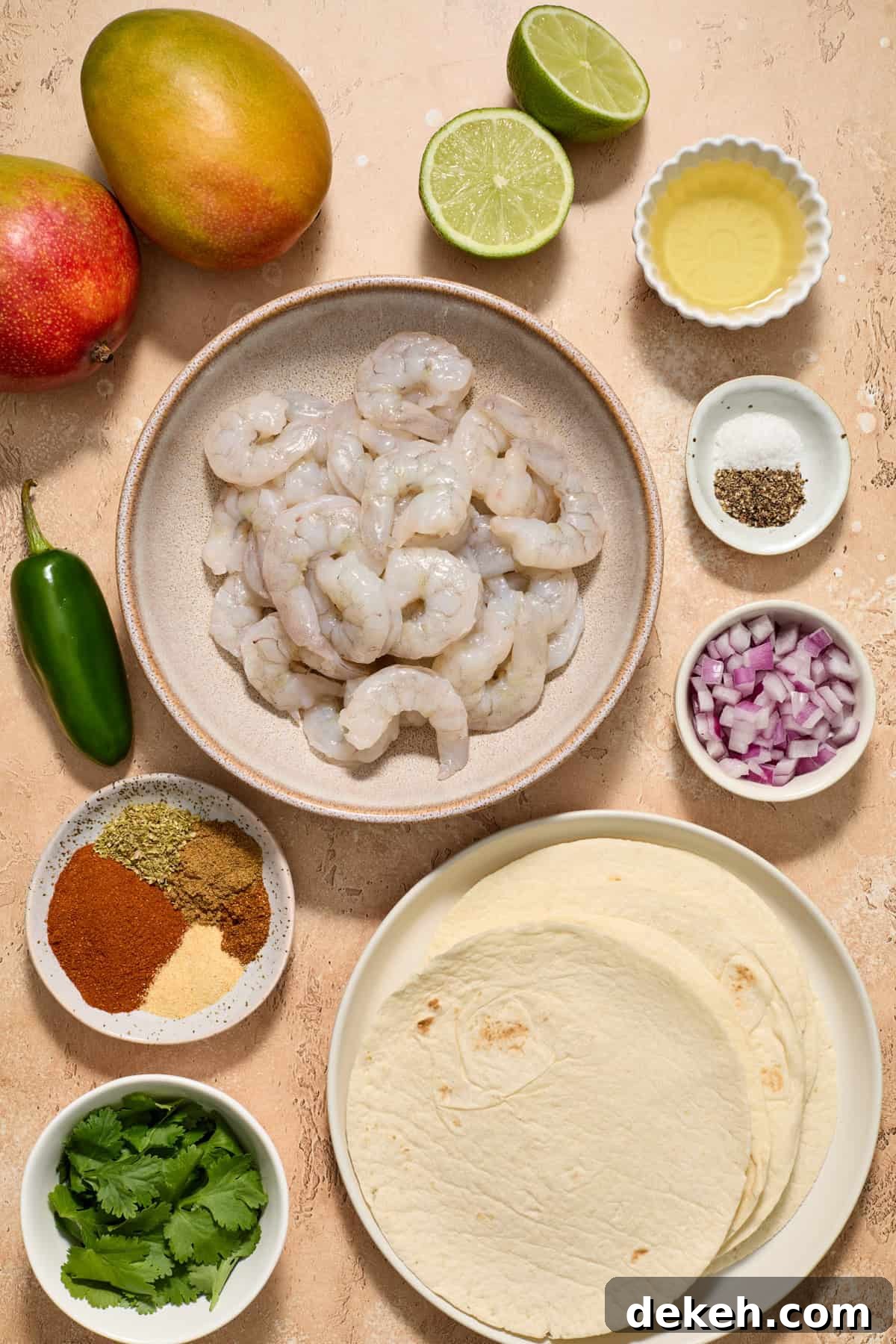 Raw shrimp, mango, spices, tortillas, limes and other ingredients arranged on surface.