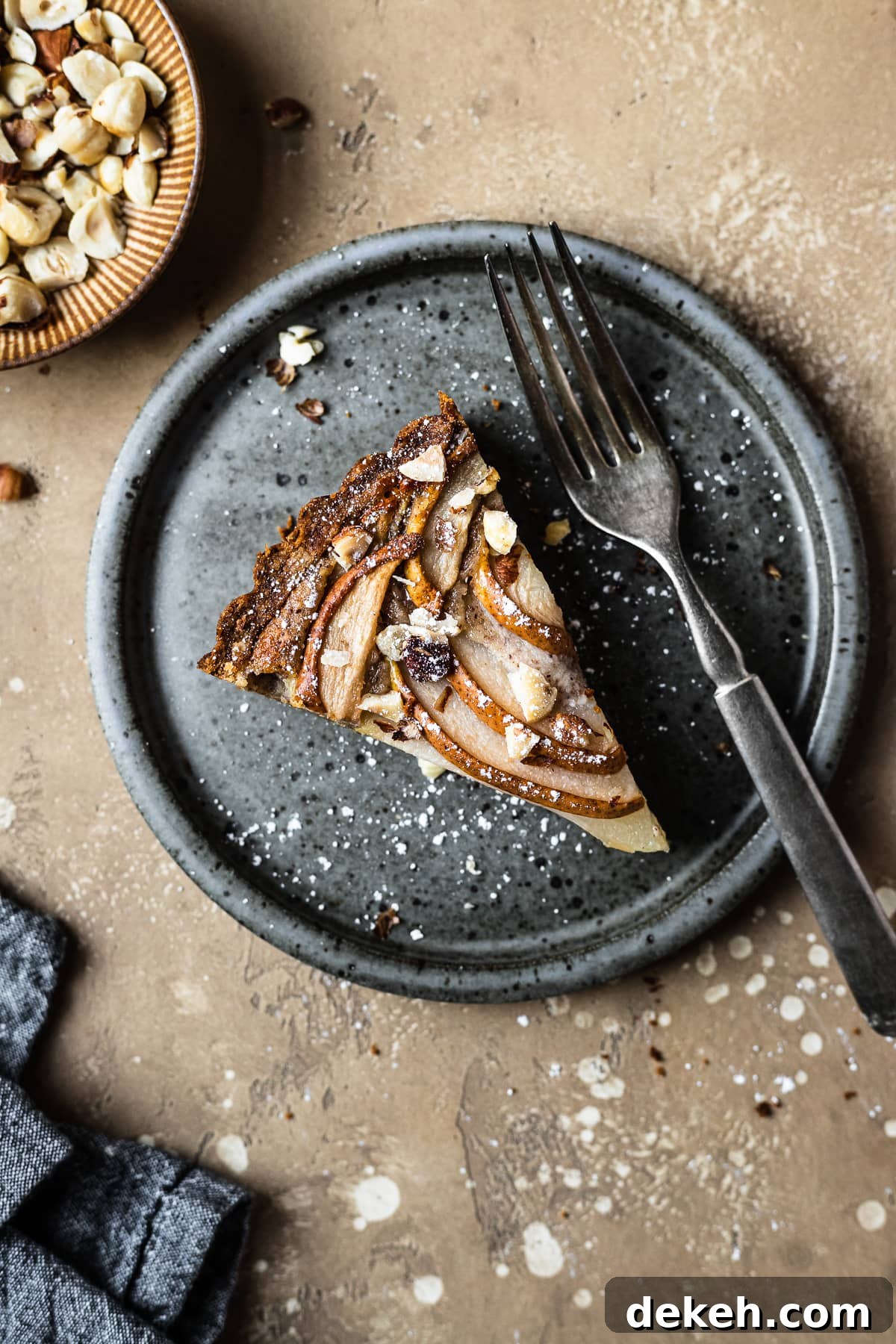 Pear hazelnut tart slice on a speckled grey blue ceramic plate. A vintage fork rests on the right side of the plate. The plate is on a warm tan textured stone background. A small bit of a textured grey napkin peeks out from the bottom left corner, and a small view of a bowl of chopped hazelnuts is visible at top left.