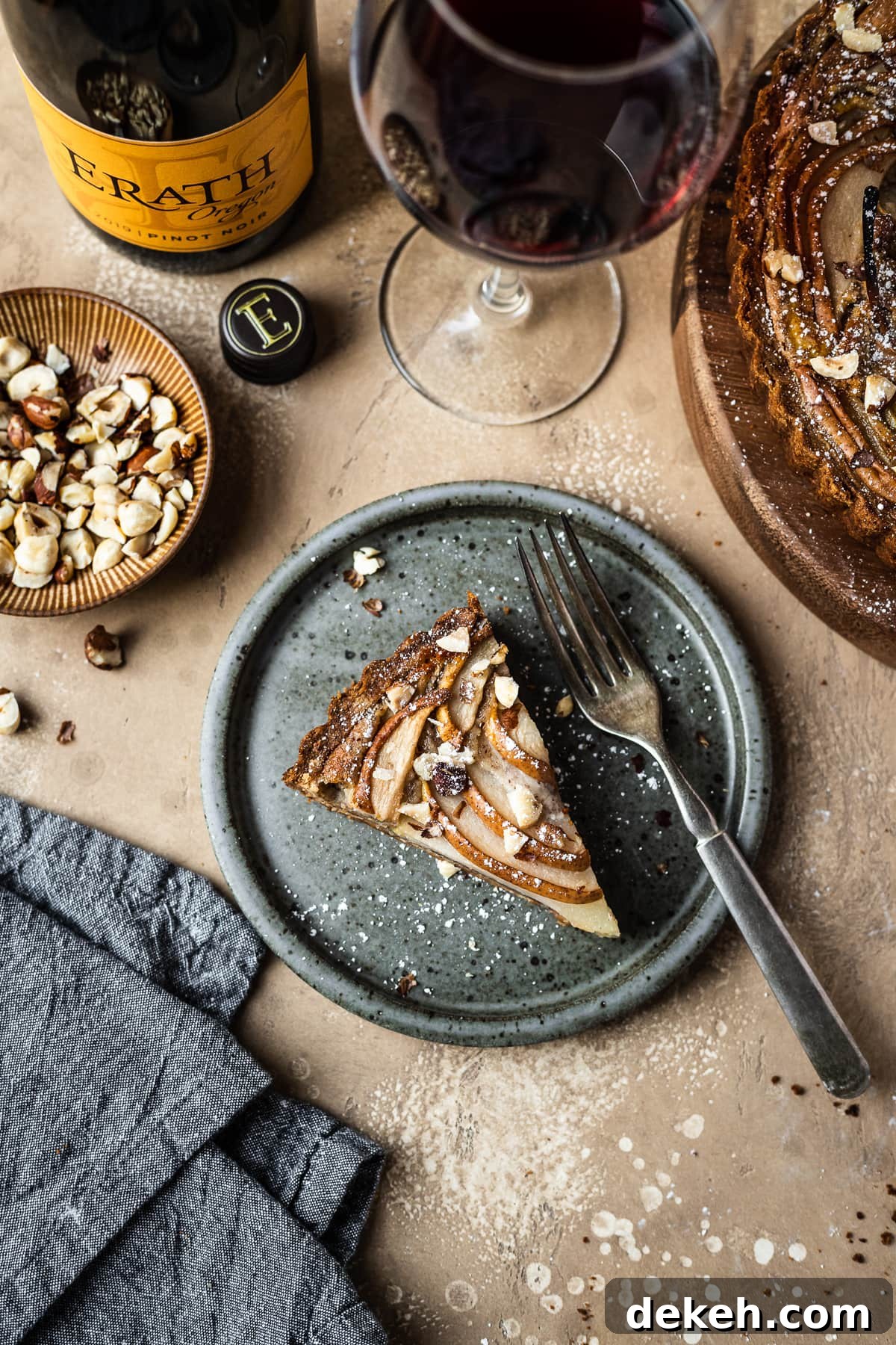 Pear hazelnut tart slice on a speckled grey blue ceramic plate. A vintage fork rests on the right side of the plate. The plate is on a warm tan textured stone background. A textured grey napkin is at the bottom left corner, and a small bowl of chopped hazelnuts is visible at top left along with a wine bottle and a glass of red wine at top. The remainder of the tart peeks out at top right.