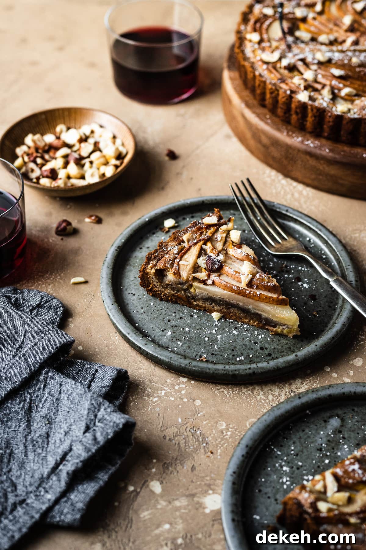 An angled view of two slices of tart on speckled grey blue ceramic plates. Nearby is the remainder of the tart, a small bowl of hazelnuts, and two stemless wine glasses filled with red wine. A grey textured linen napkin is at bottom left. The background of the image is a warm tan speckled stone.