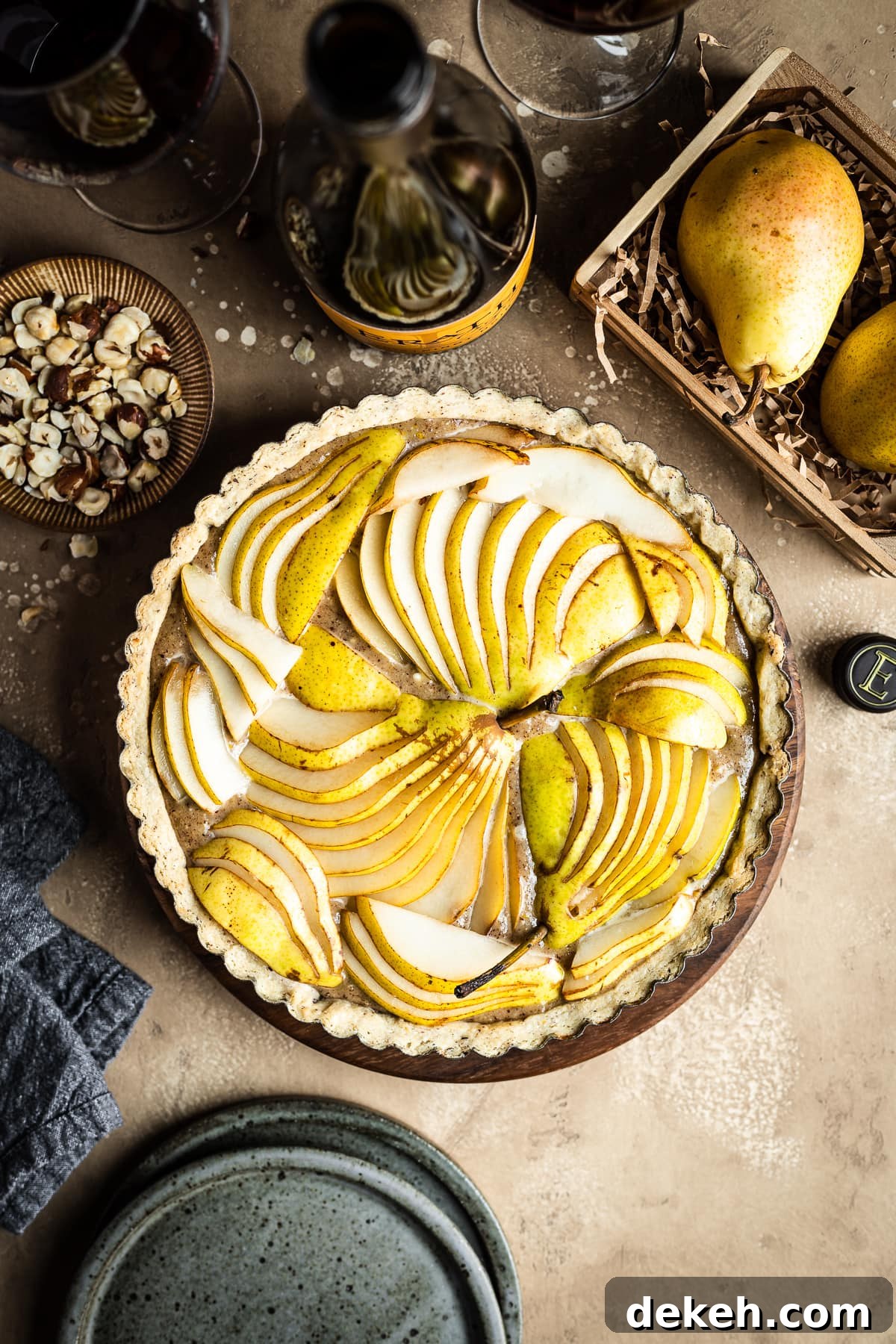 An unbaked pear hazelnut tart in a round pan is at the center of the image. Surrounding the tart are speckled blue ceramic plates, a bowl of chopped hazelnuts, pears in a wooden crate, a linen napkin, and a bottle of red wine with two filled wine glasses. Everything rests on a textured tan background.