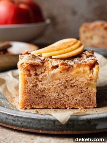 A slice of moist apple cake on a ceramic plate with three thin slices of fresh apple on top.