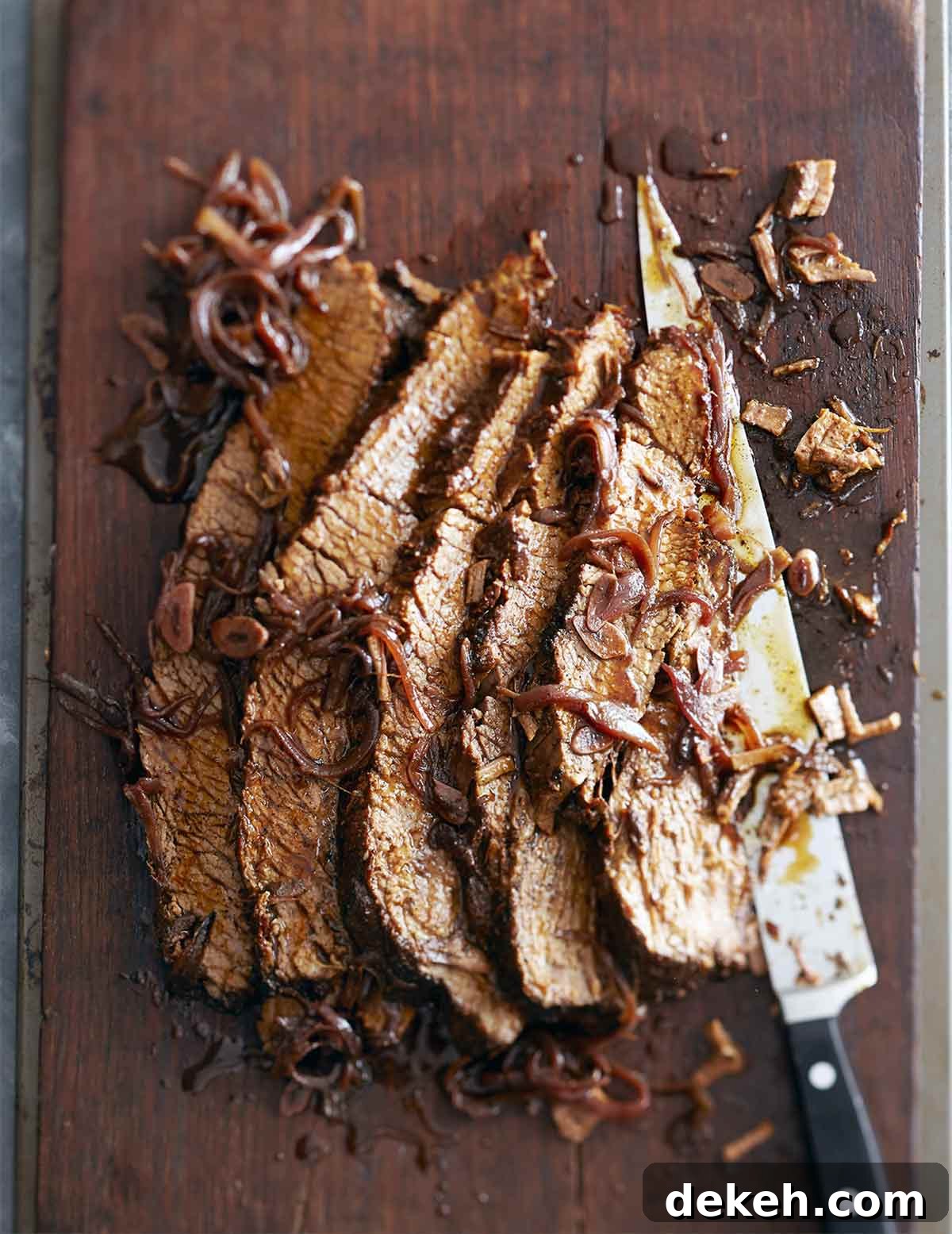 A sliced piece of braised brisket with red wine and honey laying on a cutting board with a large knife and covered with braised onions.