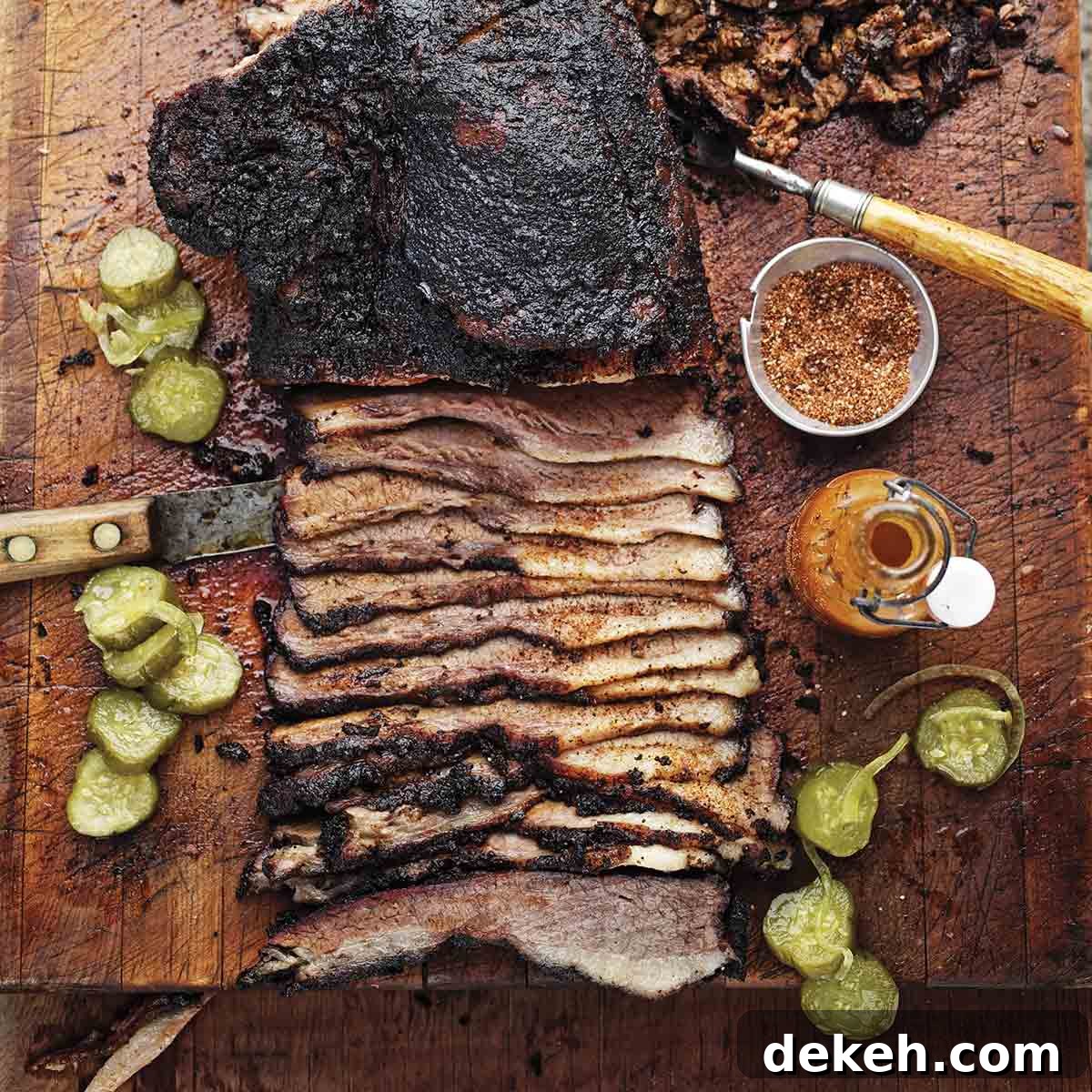A partially sliced barbecue beef brisket on a cutting board with spice rub, hot sauce, and pickle slices scattered around it.