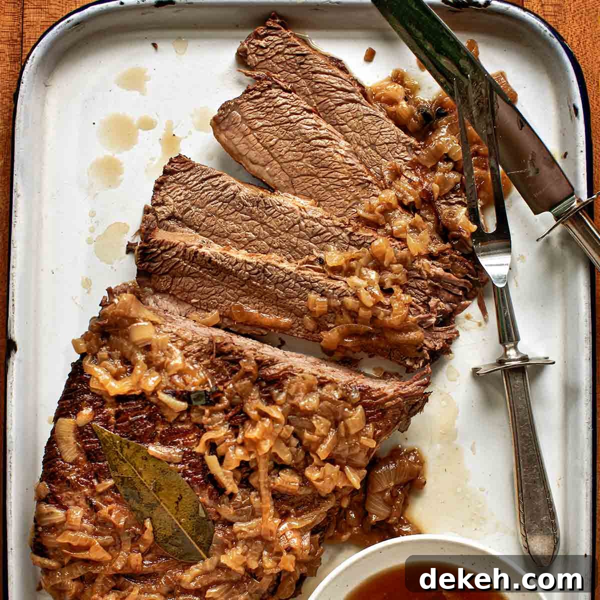 An enamel baking dish holding a sliced oven brisket topped with onions with jus on the side.