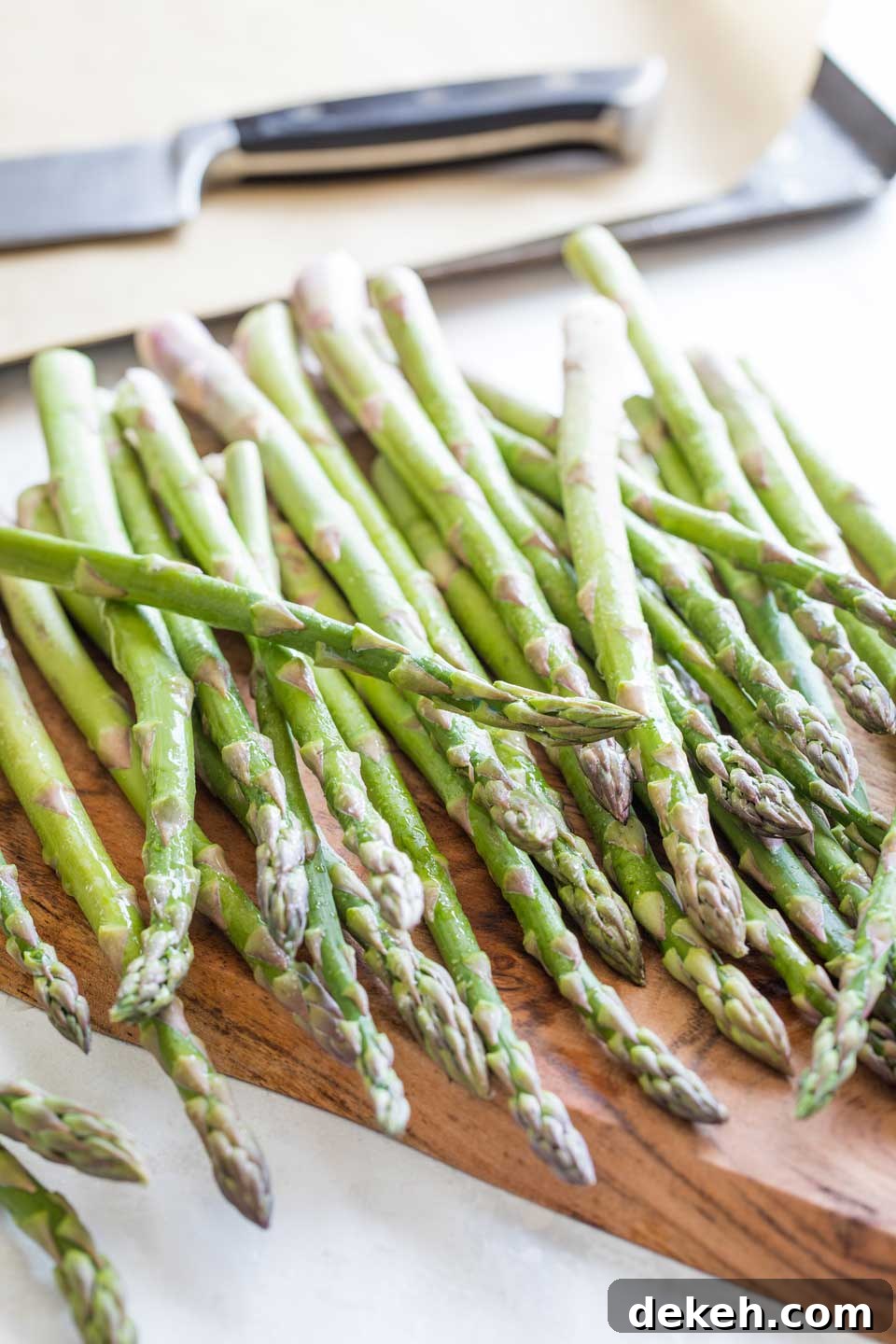 A pound of asparagus, rinsed and rolled across a wooden cutting board with a roasting pan and knife in the background.