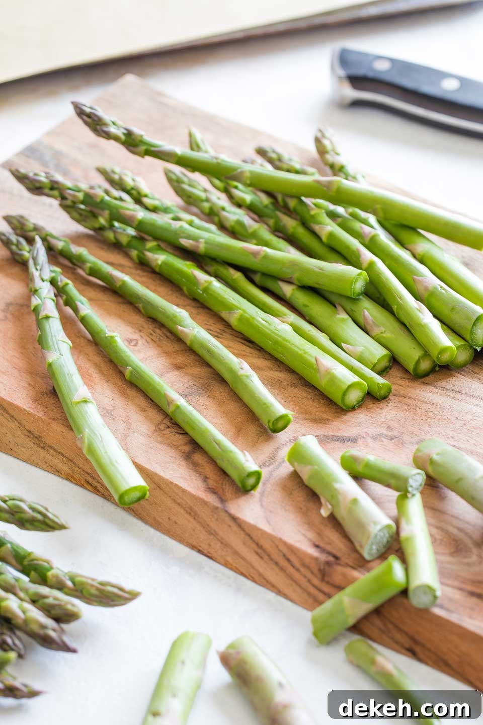 13 asparagus stalks on a wooden cutting board, with the bottom inch or two just sliced off and the discarded ends laying about nearby.
