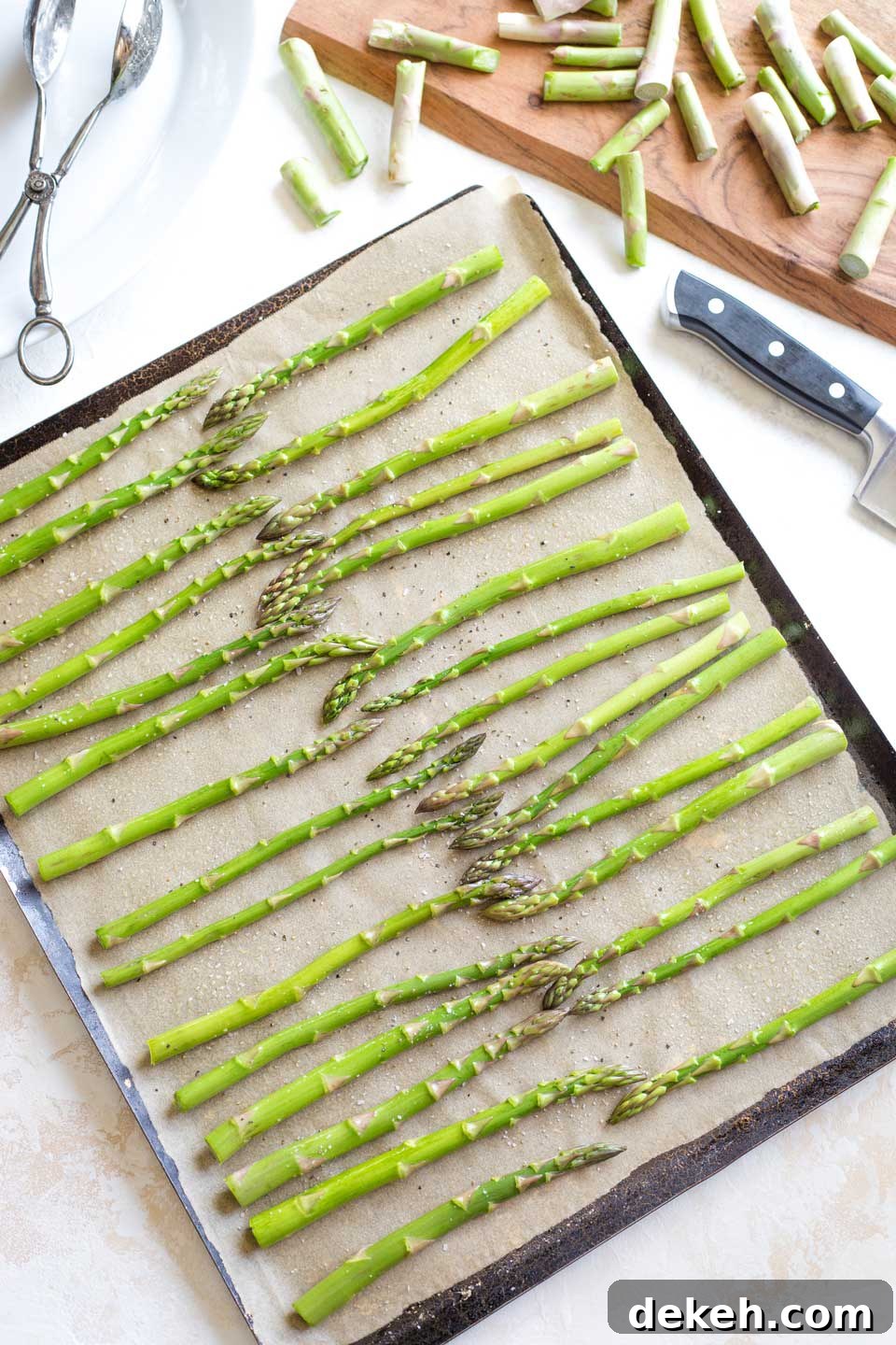 Overhead of a parchment-lined sheet pan ready for the oven, with raw asparagus arranged in a single layer and the discarded ends laying on a nearby cutting board.