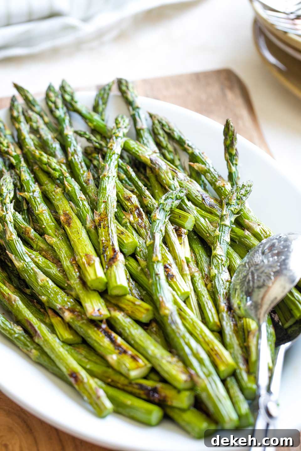 A 3/4 angle shot from the bottom of the asparagus stalks, with the finished stalks piled on a white platter (next to serving tongs) so you can see lots of the brown, roasted spots.