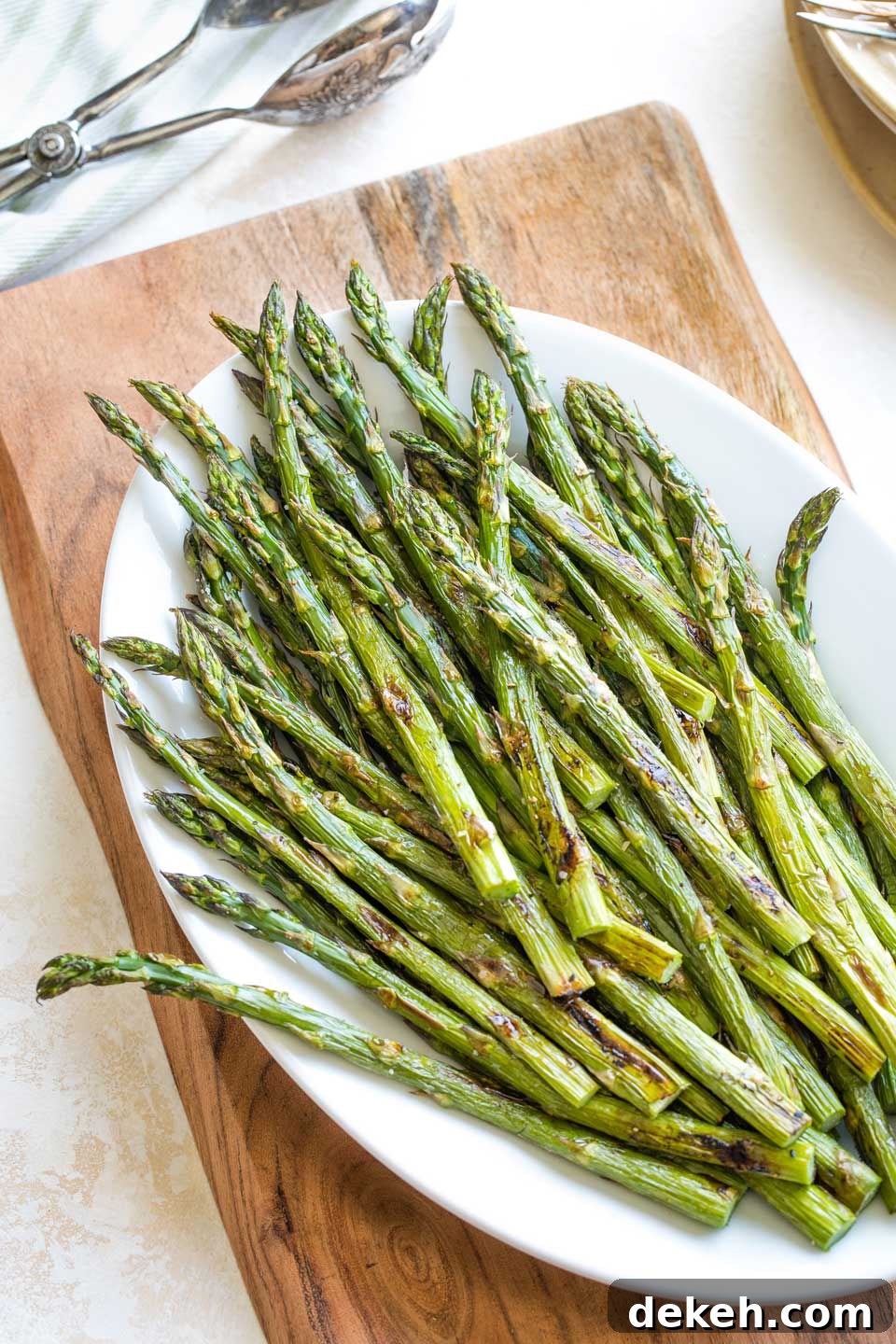 Overhead of the full white serving platter loaded with a serving of oven roasted asparagus, with serving tongs and a stack of plates and forks waiting nearby.