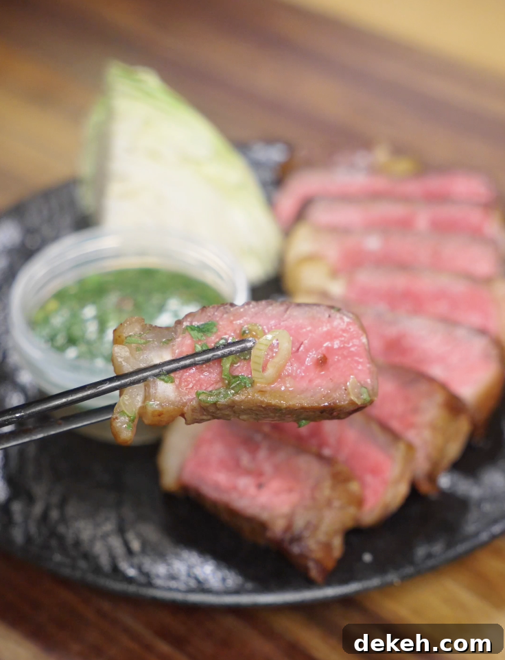 Close-up of a chopsticks bite of medium-rare Crying Tiger Steak dipped in Thai-style sauce, showing juicy steak and fresh herbs.