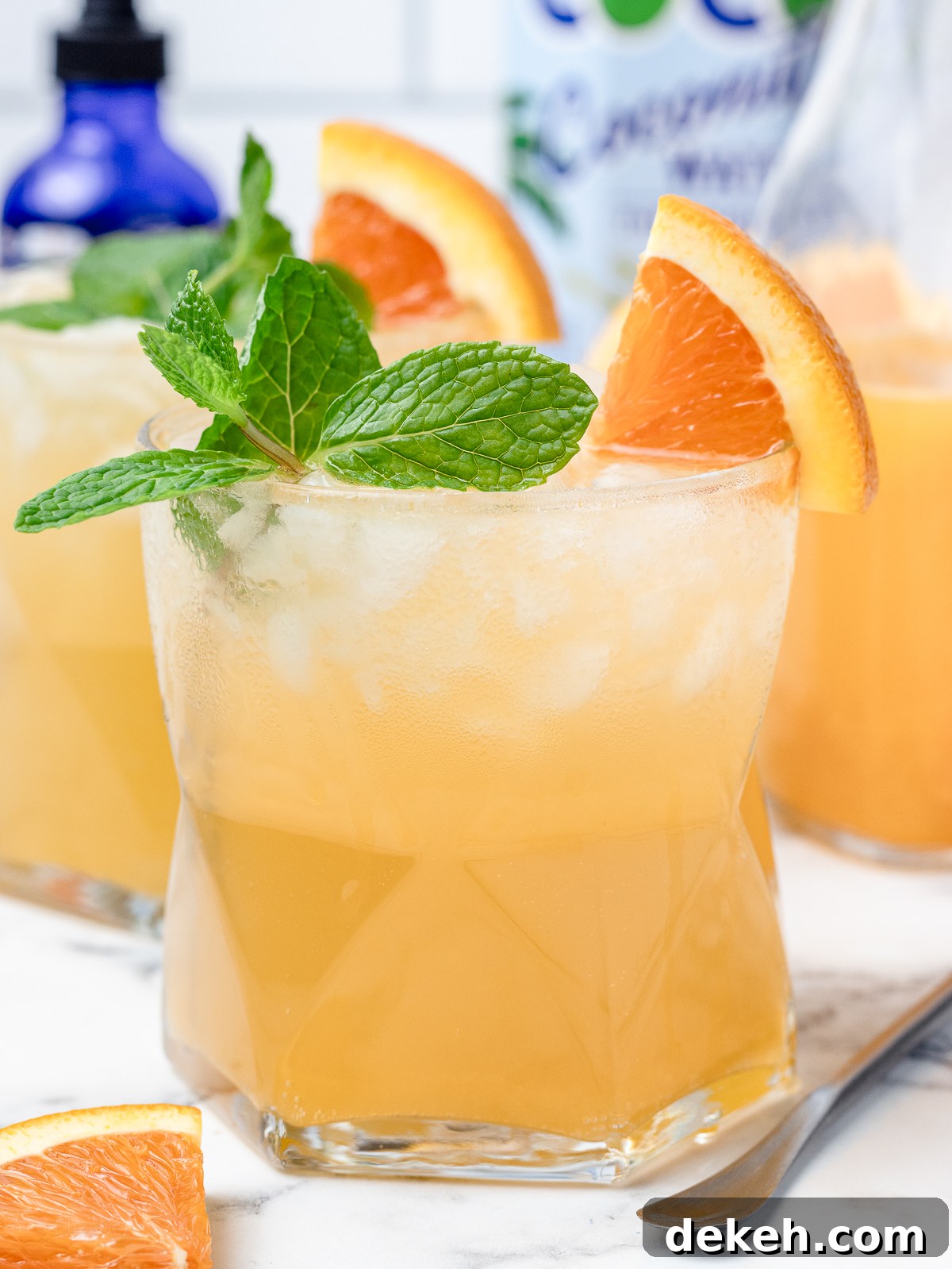 An iced Orange Coconut Water Mocktail with fresh mint and orange slices. They are a light and refreshing orange color. There is freshly squeezed orange juice, alcohol free orange bitters, and coconut water in the background.