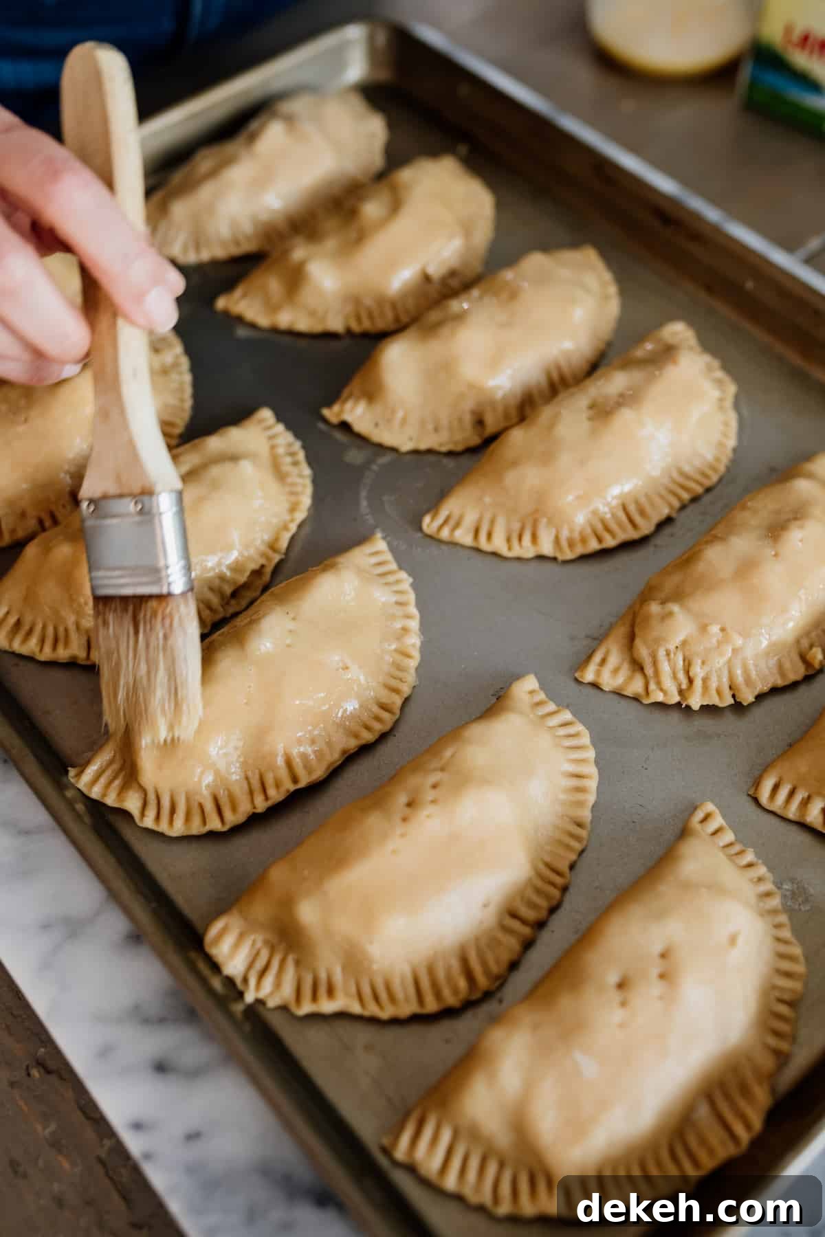 brushing egg wash on empanadas on a baking sheet.