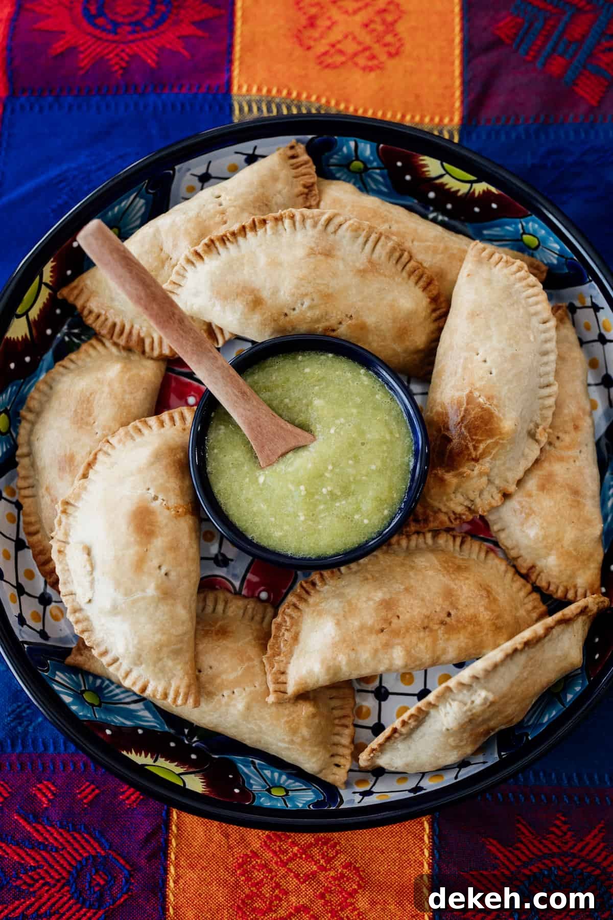 empanadas displayed on a Mexican talavera serving tray with a dish of salsa verde on top of a colorful Mexican tablecloth.