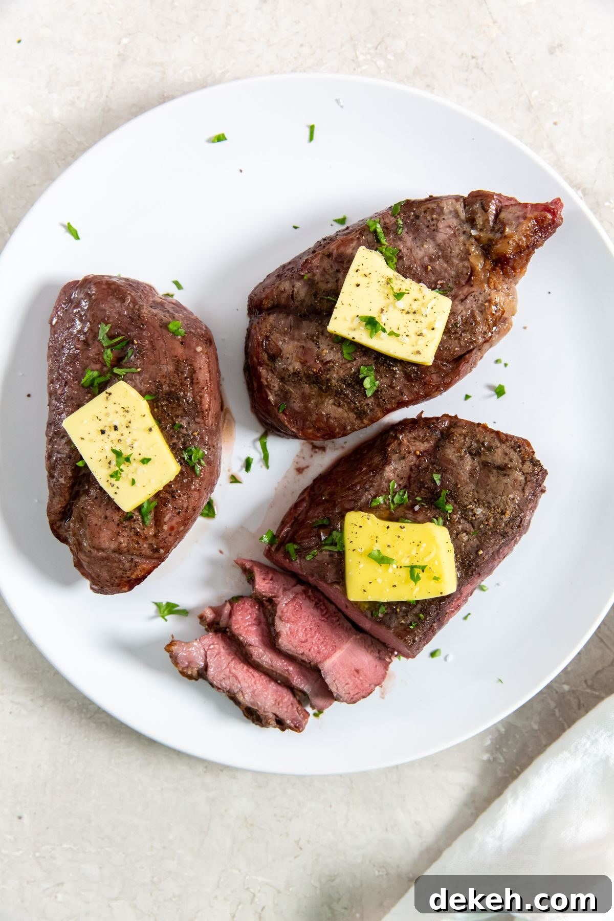 Three cooked and sliced petite sirloin steak topped with butter and herbs on a white plate.
