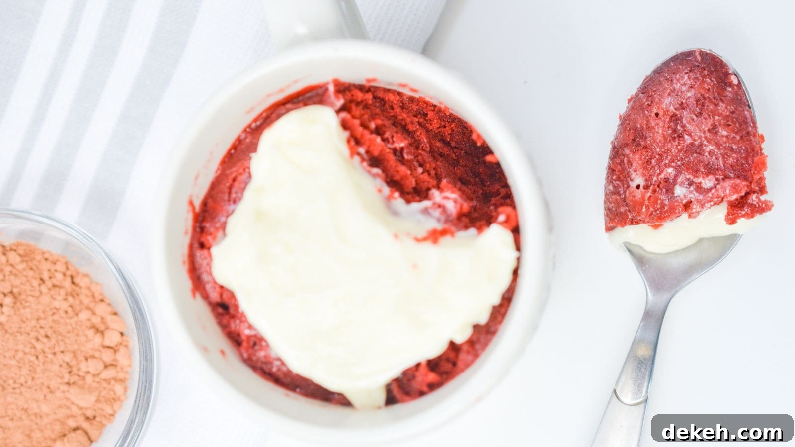 Red velvet cake with a layer of cream cheese frosting in a white ramekin, next to a spoon with a bite taken out, on a white background.