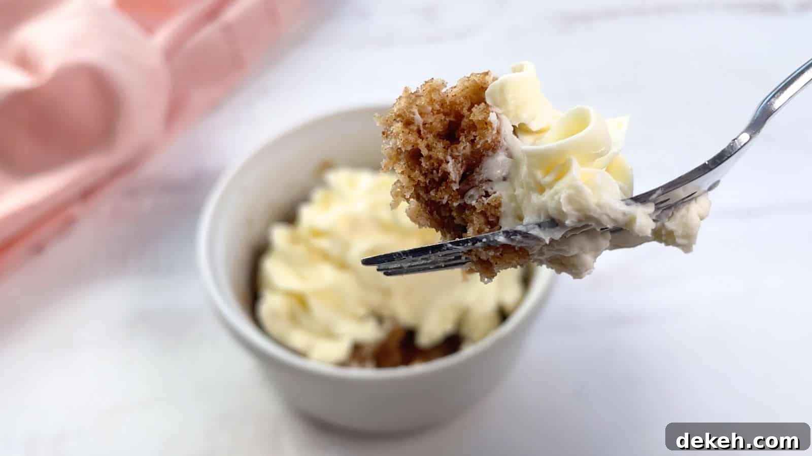 A fork holding a piece of cake with cream cheese frosting over a frosted cake mix mug cake on a table.