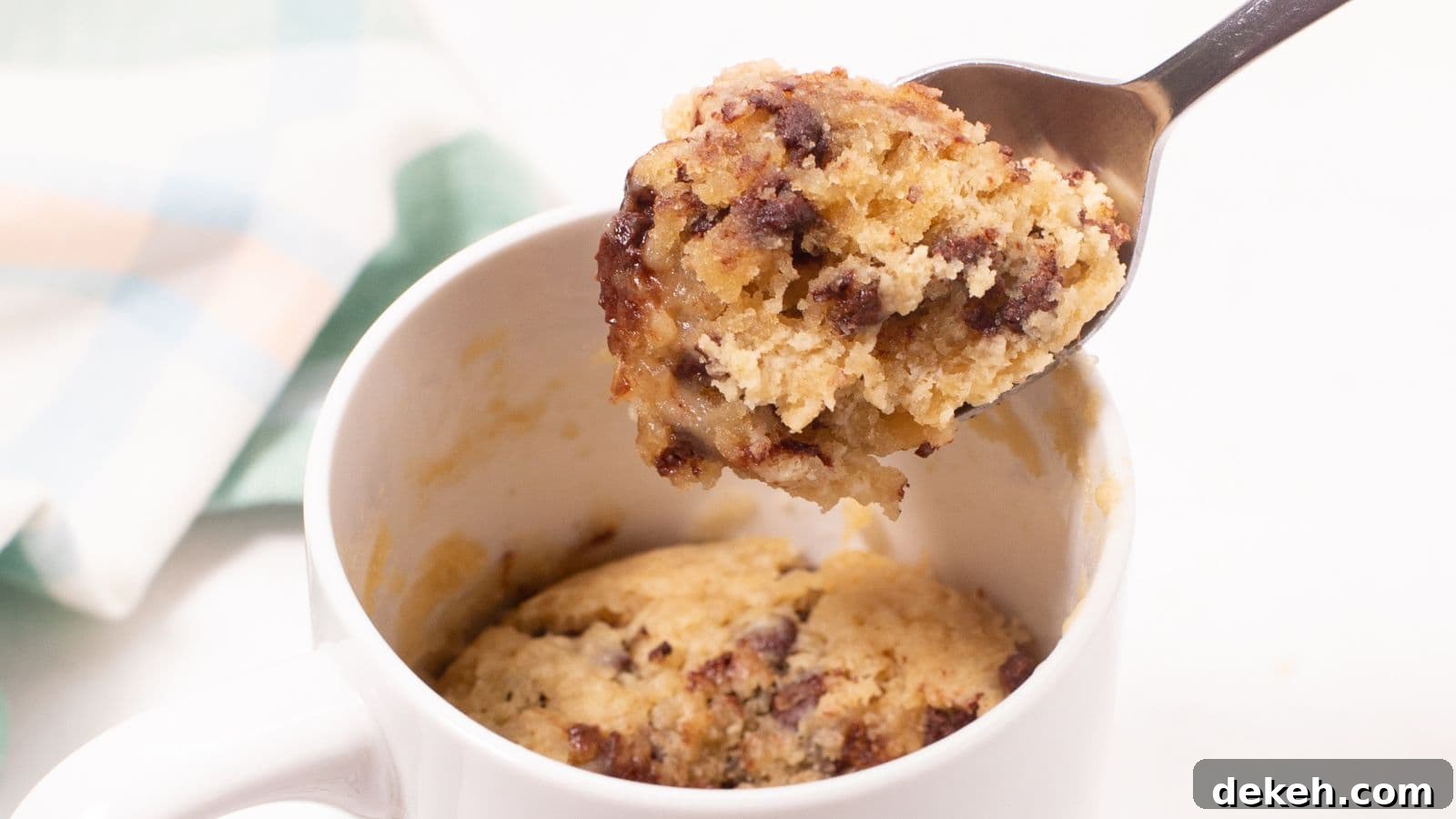 A spoon lifting a bite from a chocolate chip mug cake in a white mug.
