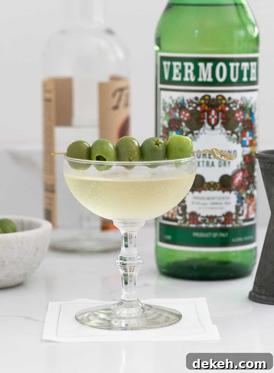 Dirty martini in a coupe with olives and a bottle of Vermouth in the back.