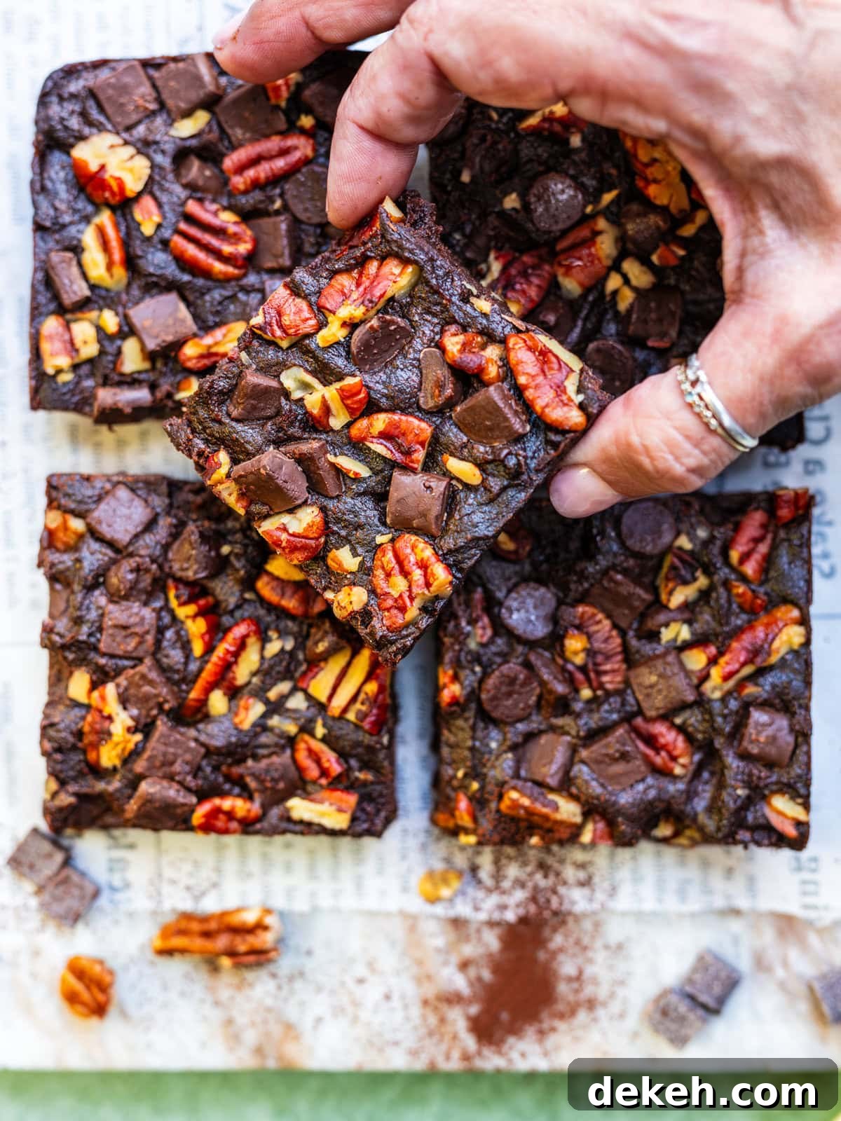 A hand picks up a square of sweet potato brownie topped with pecans and chocolate chunks, with more brownies arranged on newspaper below. Cocoa powder, pecans, and chocolate pieces are scattered nearby.
