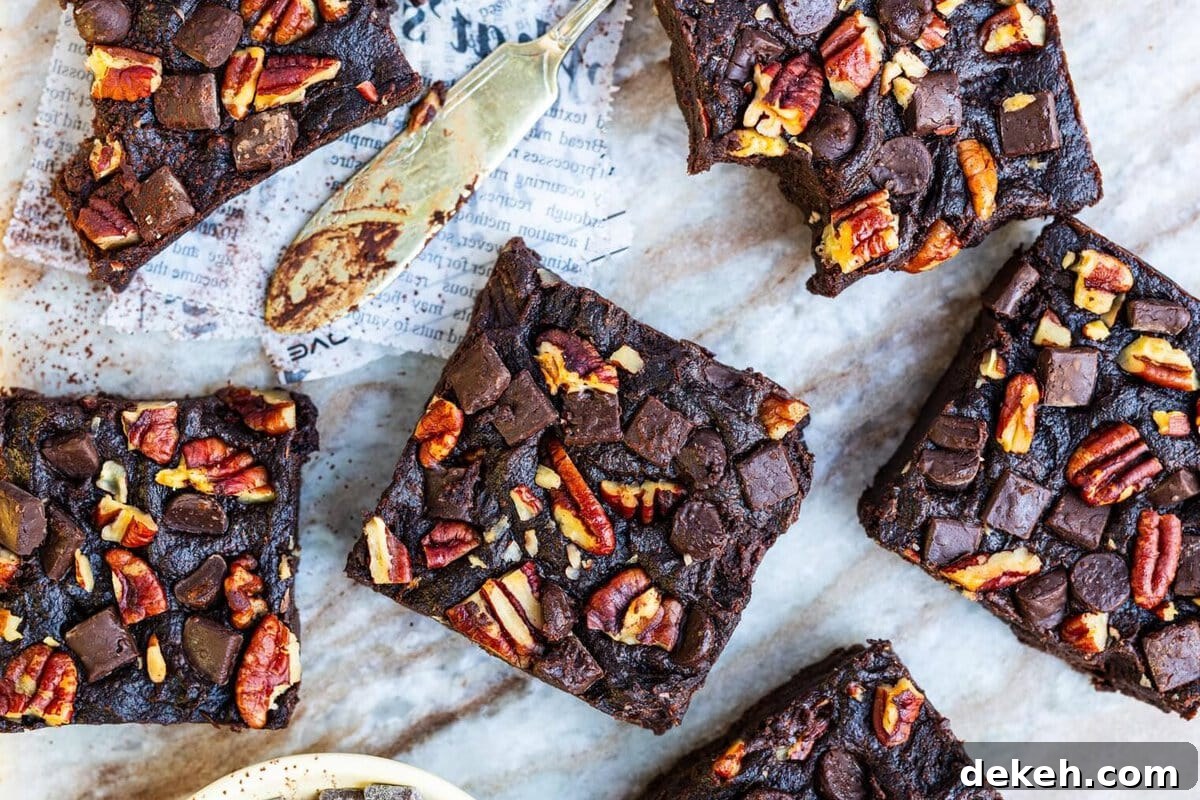 Five squares of rich, chocolate brownies topped with pecans and chocolate chunks are arranged on a marble surface, with a butter knife and a small bowl of chocolate pieces nearby.