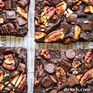 Four sweet potato brownies topped with pecans, chocolate chunks, and chocolate chips are arranged closely together on a sheet of newspaper. The brownies have a rich, fudgy texture and are generously studded with toppings.
