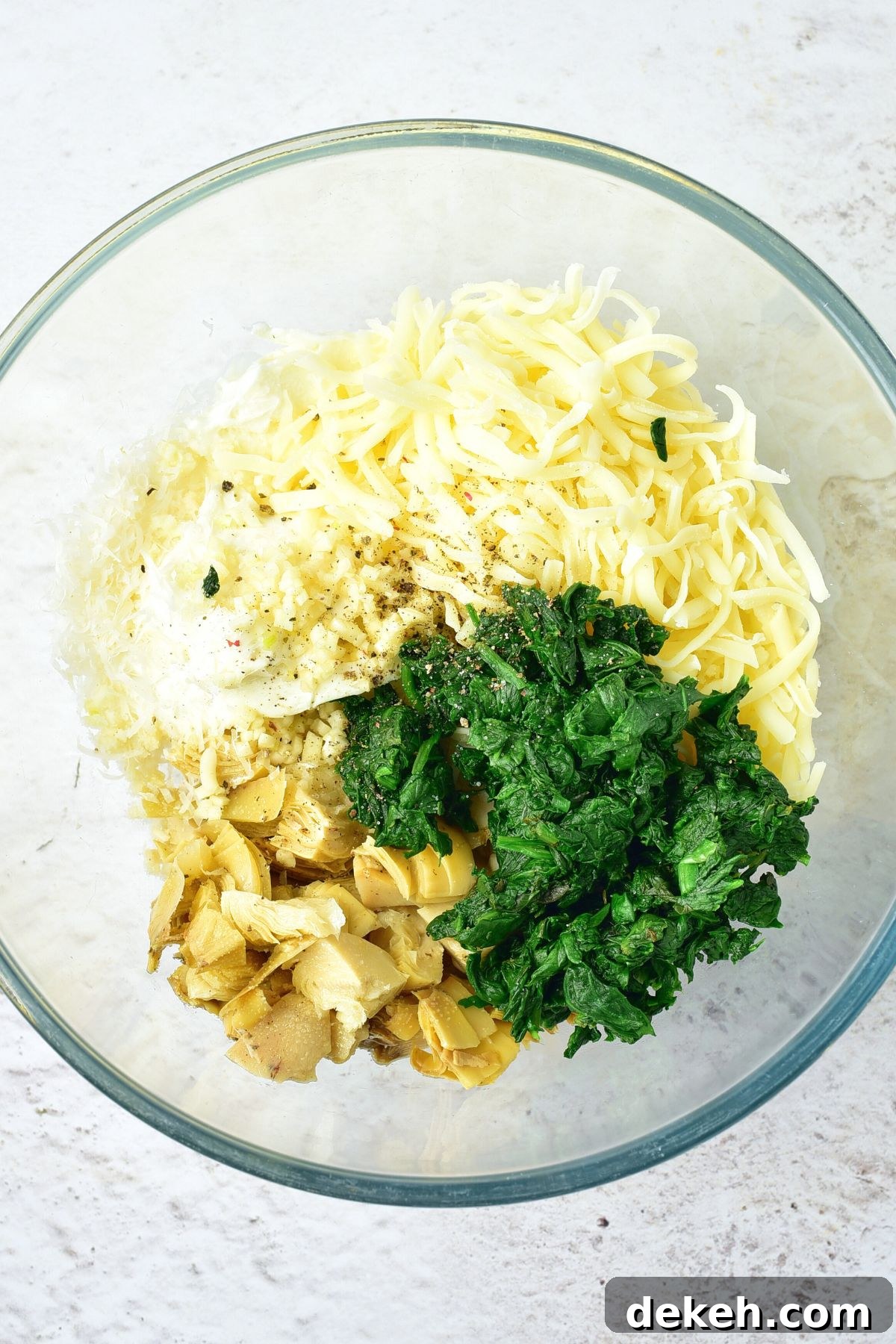 Shredded cheese, wilted spinach, and chopped artichokes in glass bowl