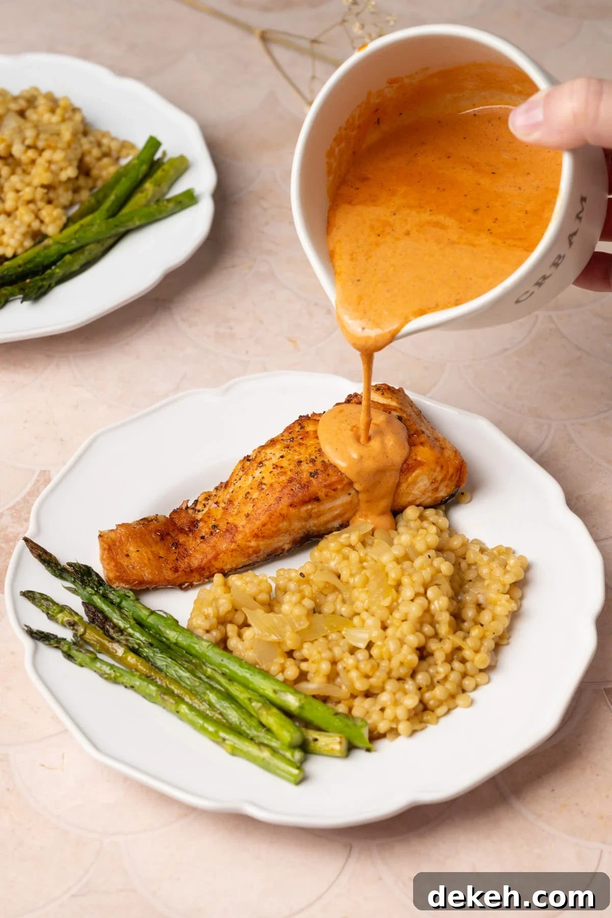 Pouring creamy Cajun sauce over salmon on a dinner plate with couscous and asparagus. 
