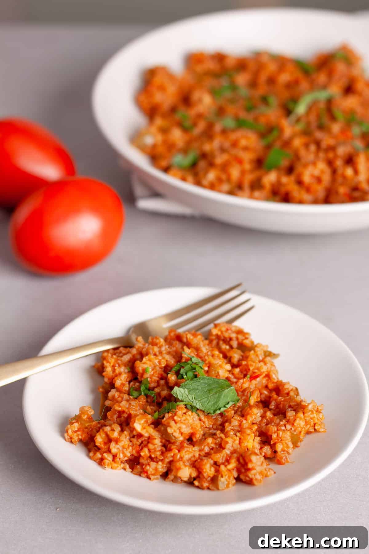 A serving of bulgur pilaf topped with fresh herbs on a small plate with a larger platter behind.