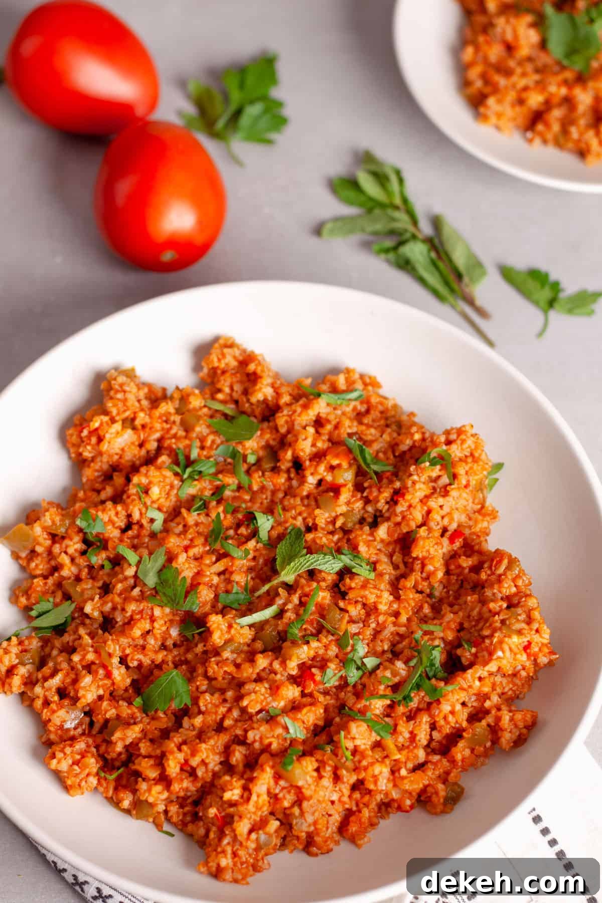 A platter of bulgur pilaf on a gray table, topped with fresh herbs and tomatoes to the side.