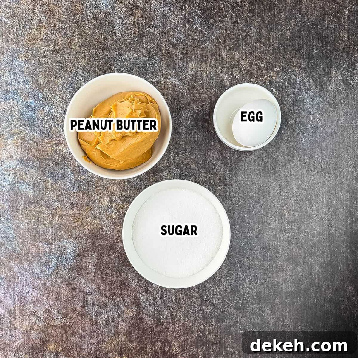 Three ingredients for air fryer peanut butter cookies laid out and labeled: bowl of creamy peanut butter, granulated sugar, and egg.