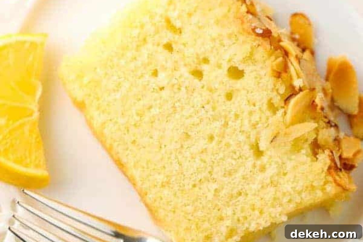 A close-up of a slice of lemon almond cake garnished with toasted almond flakes, accompanied by a wedge of lemon on a white plate.
