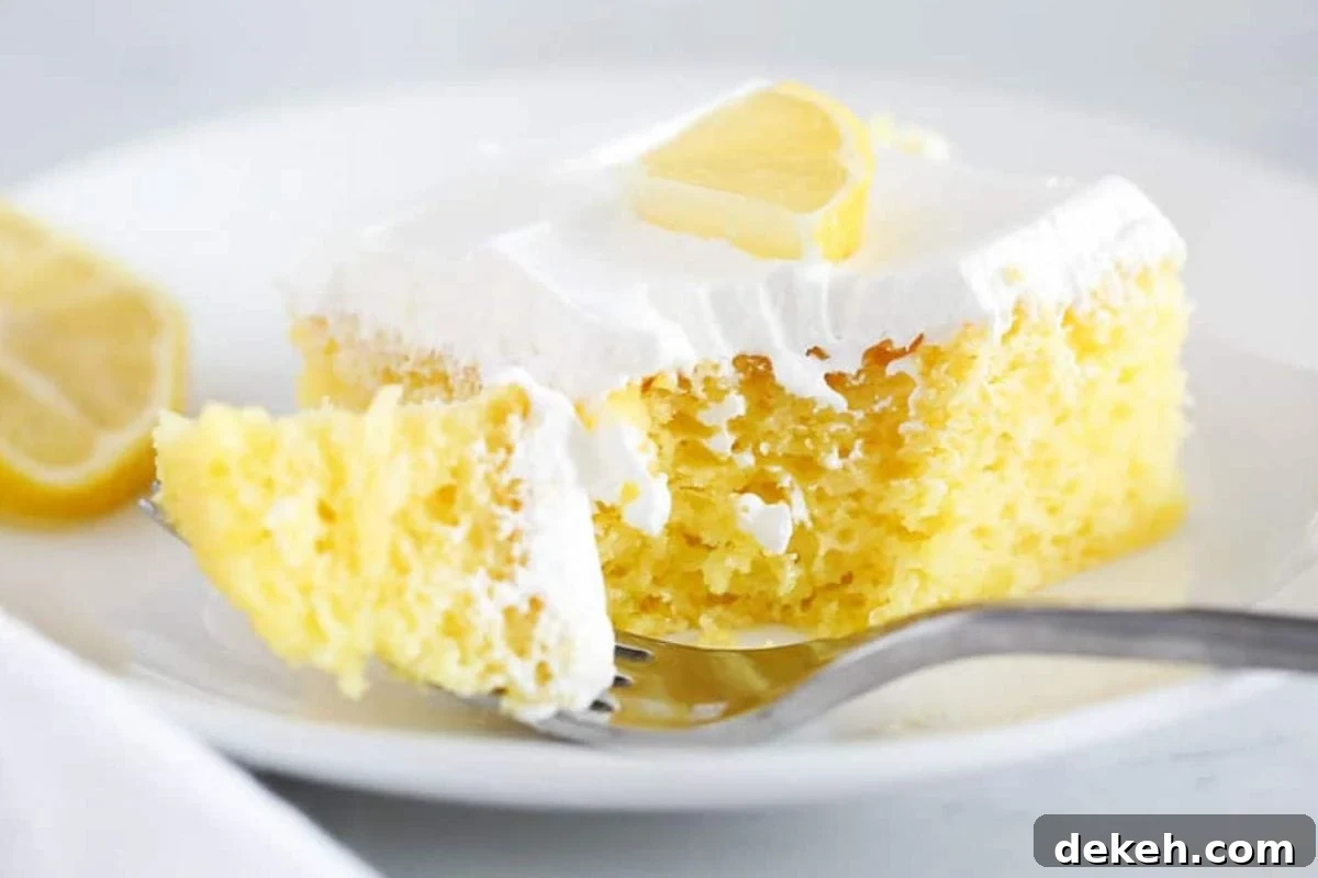 A delicious slice of lemon cake topped with creamy frosting and a slice of lemon, served on a white plate with a fork.