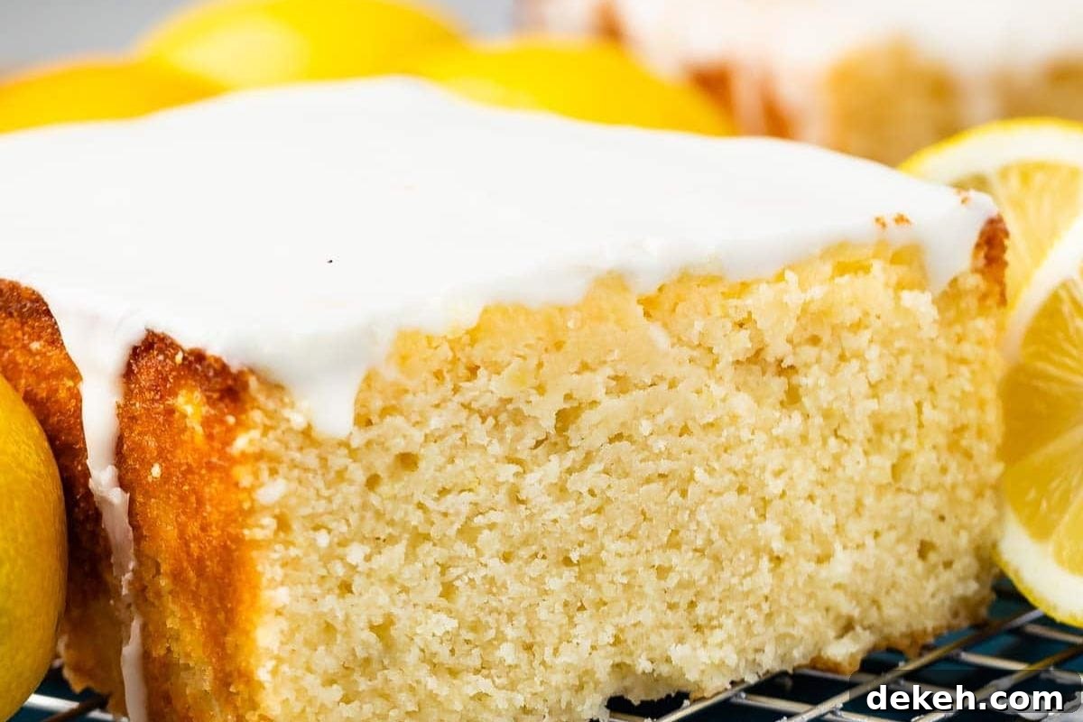 A close-up of a moist lemon cake topped with a white glaze, surrounded by fresh lemons on a cooling rack.
