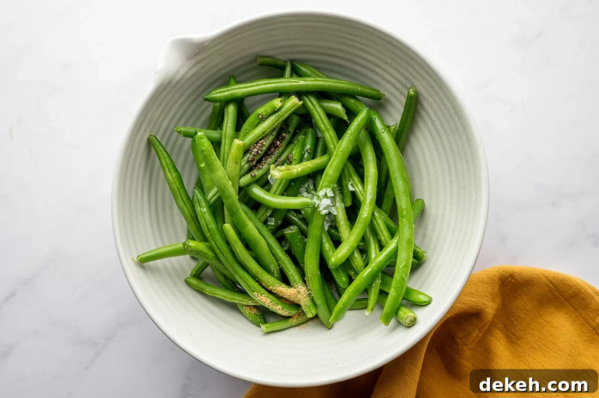 Fresh green beans in a white bowl with garlic powder, salt, and black pepper sprinkled on top.