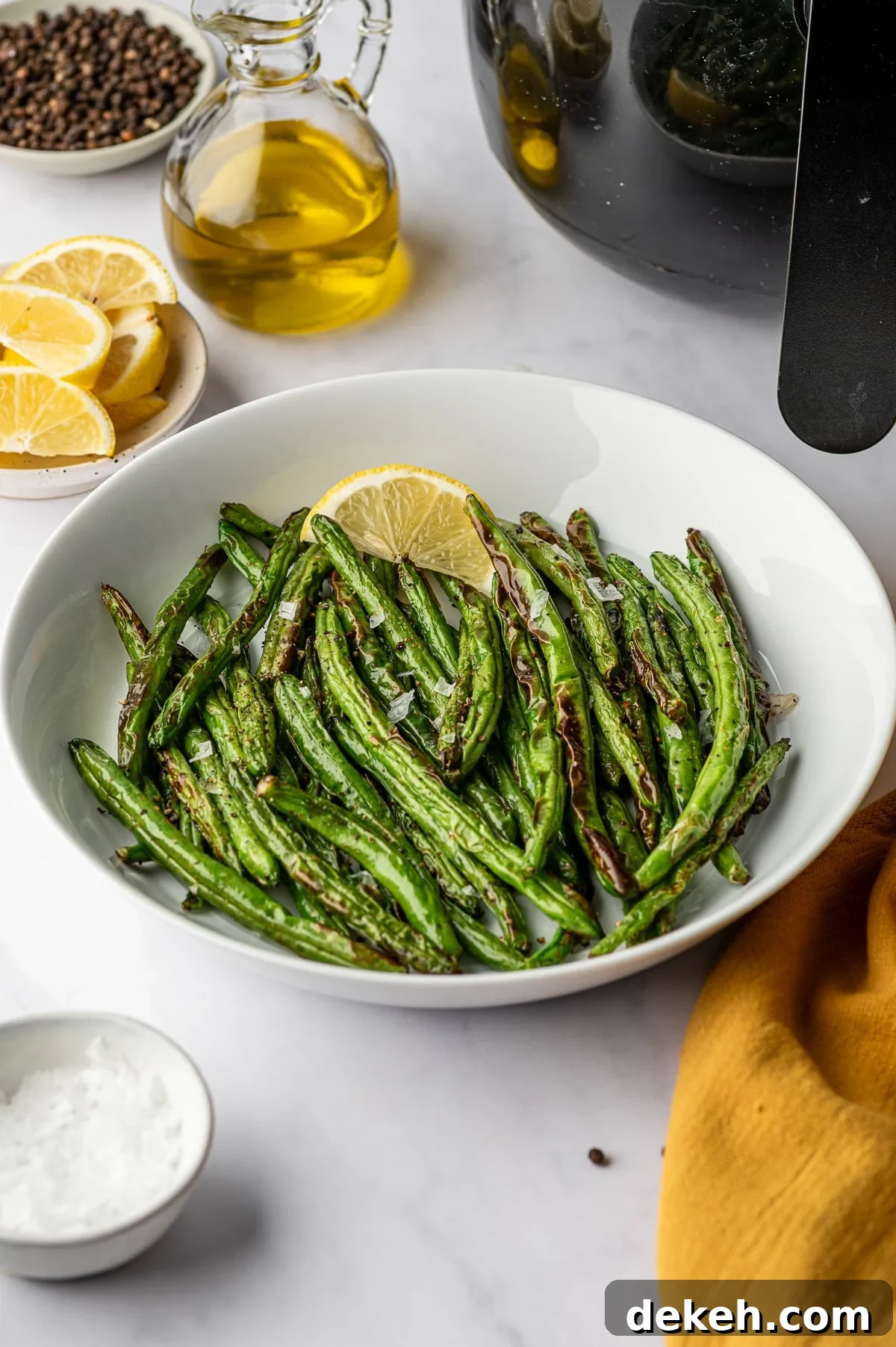 Plate of air fryer green beans with lemon slice, olive oil, and peppercorns in the background.