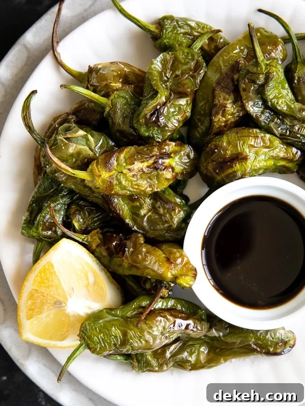White plate filled with cooked air fryer shishito peppers with a side of citrus soy sauce and a lemon wedge.