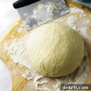 Ball of pizza dough on floured wood with dough scraper and flour bowl nearby.